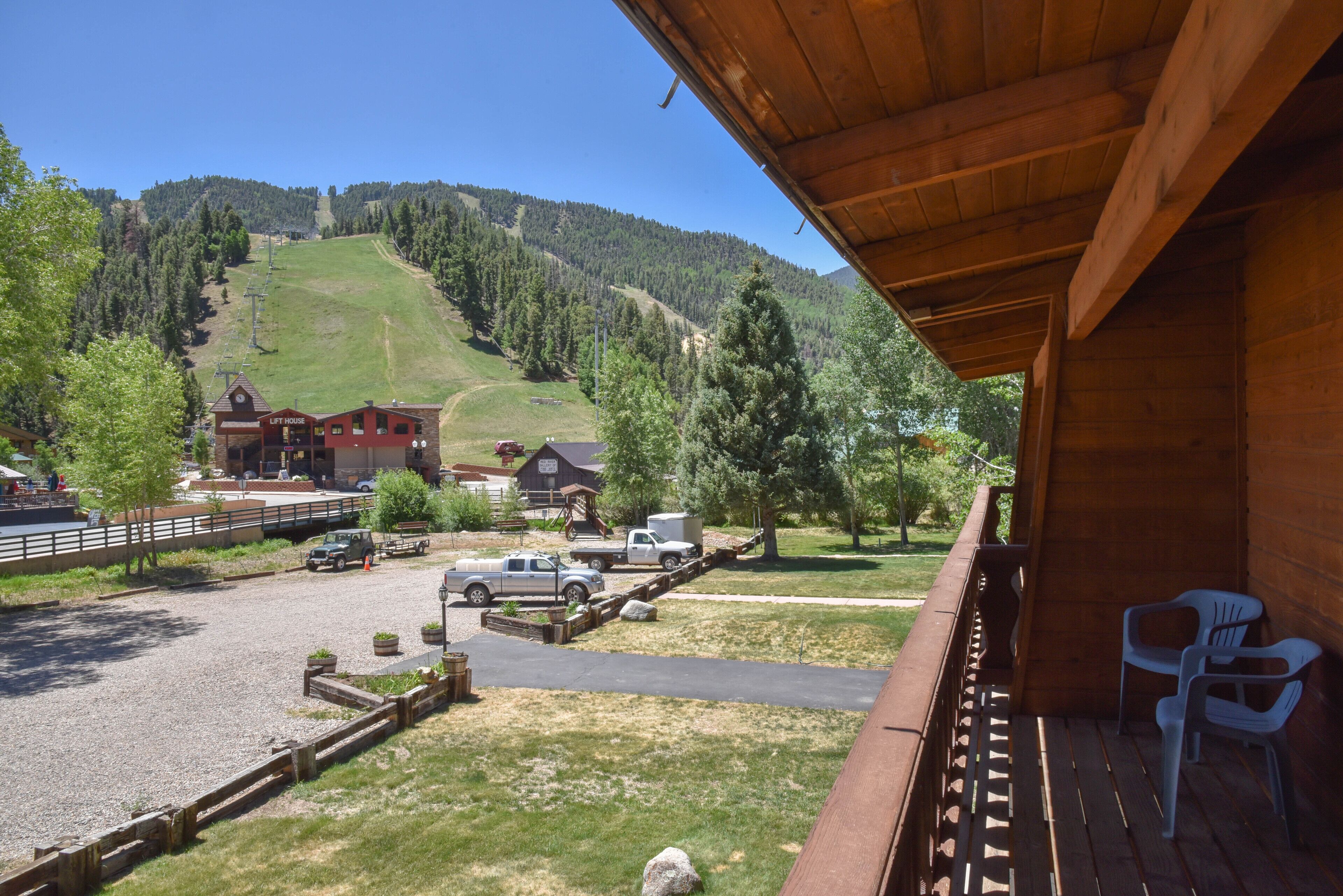 A wooden cabin with a balcony overlooking a parking area and a ski lift in the background.