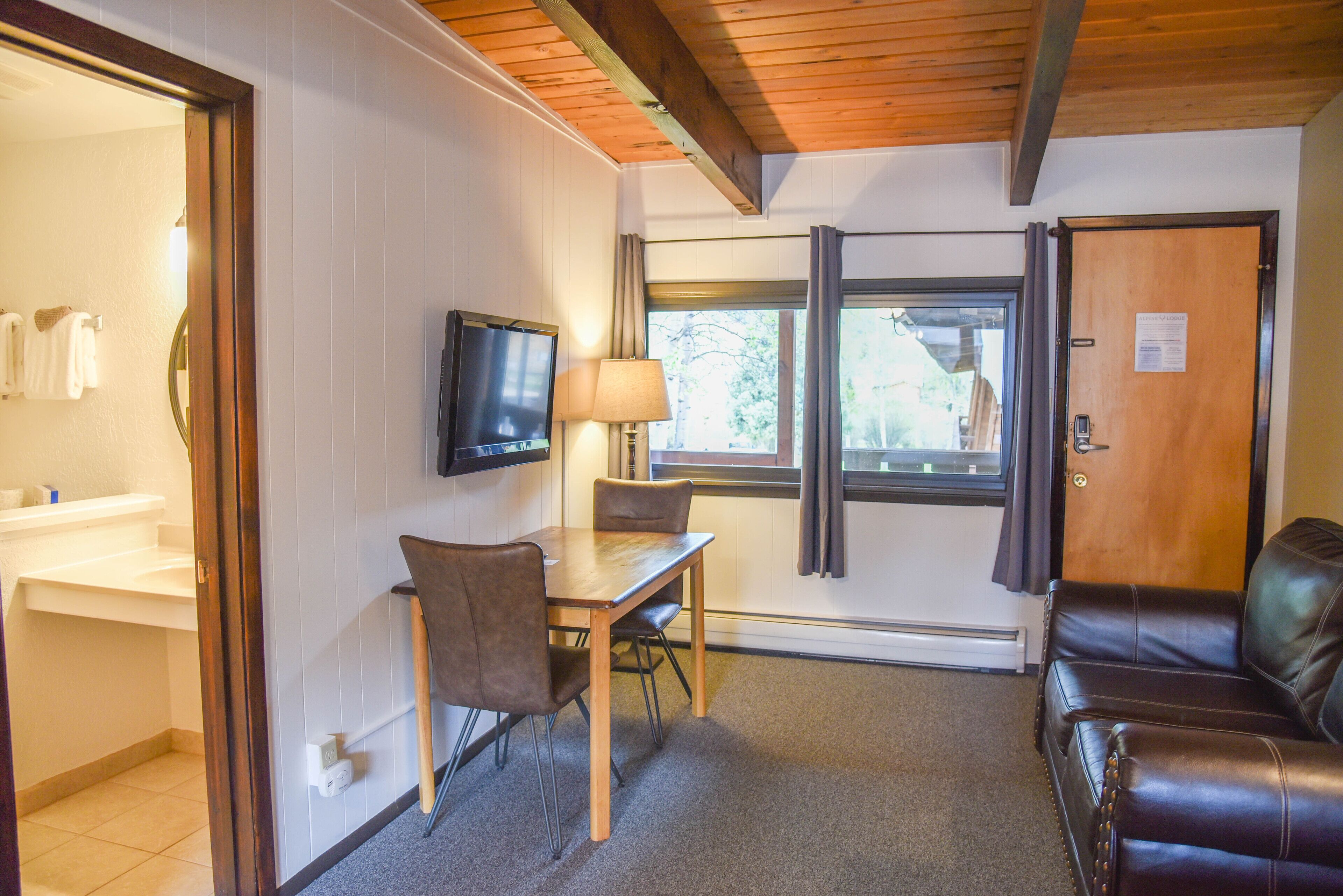 A room with a wooden ceiling, a television, a table with chairs, a sofa, and a window with curtains.