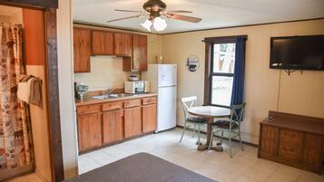 A compact kitchen with wooden cabinets, a refrigerator, a dining table with chairs, and a ceiling fan.