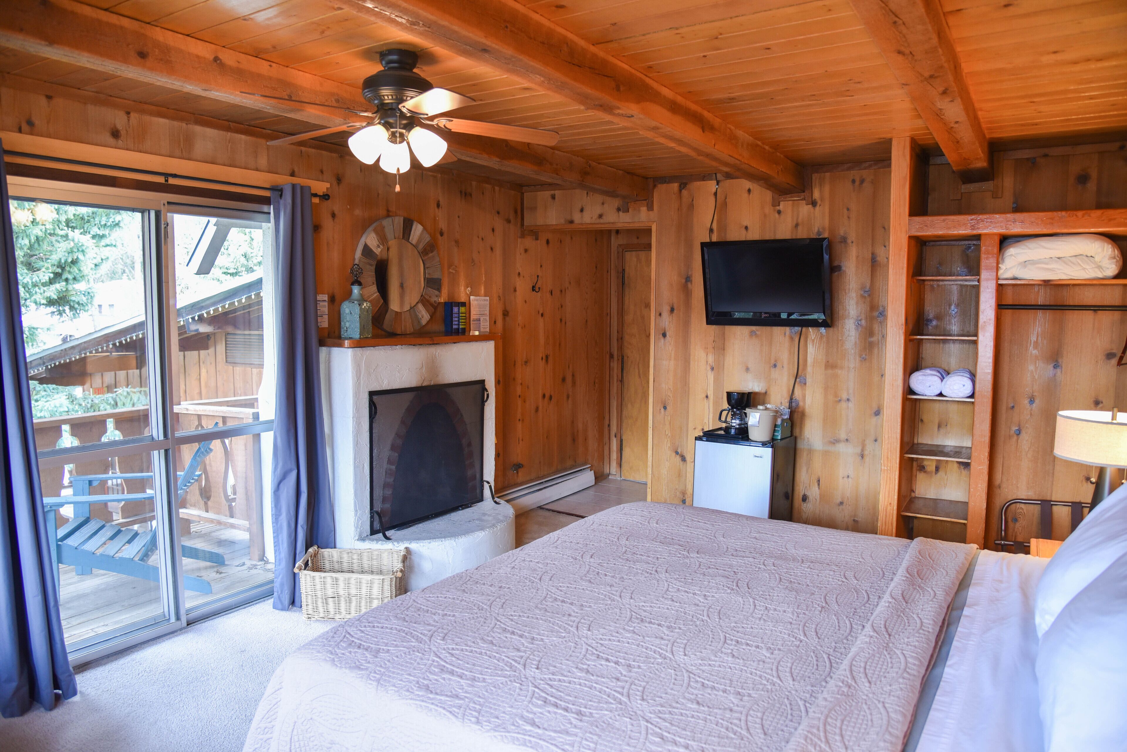 Un dormitorio de cabaña de madera con una cama grande, chimenea, televisor y balcón con mobiliario exterior.