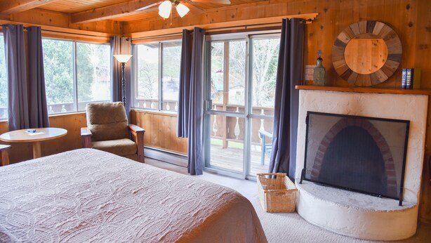 Een houten kamer met een bed, een stoel, een tafel, een open haard en grote ramen.