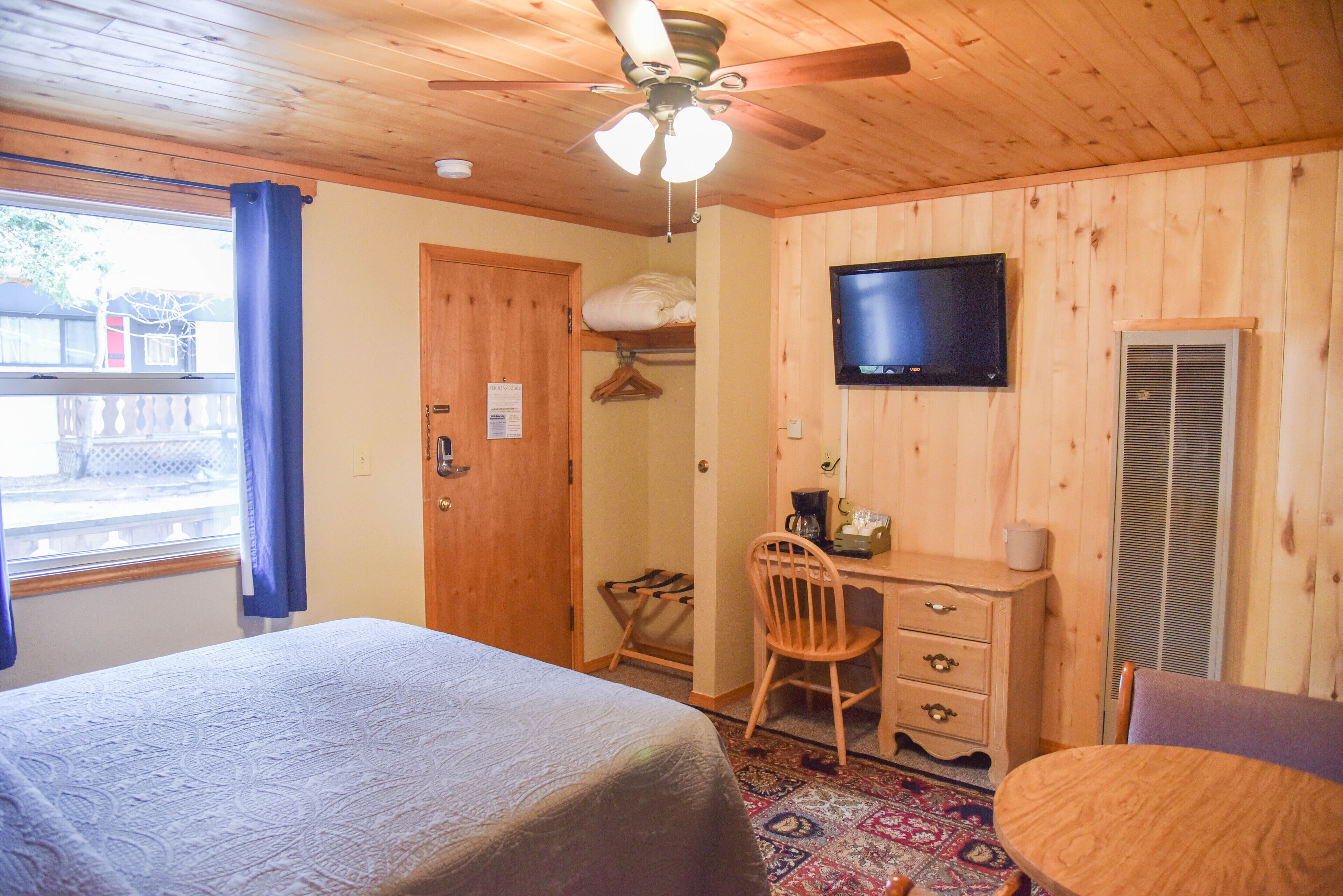 Een kamer met houten lambrisering, een bed, een bureau, een stoel, een televisie en een raam met gordijnen.