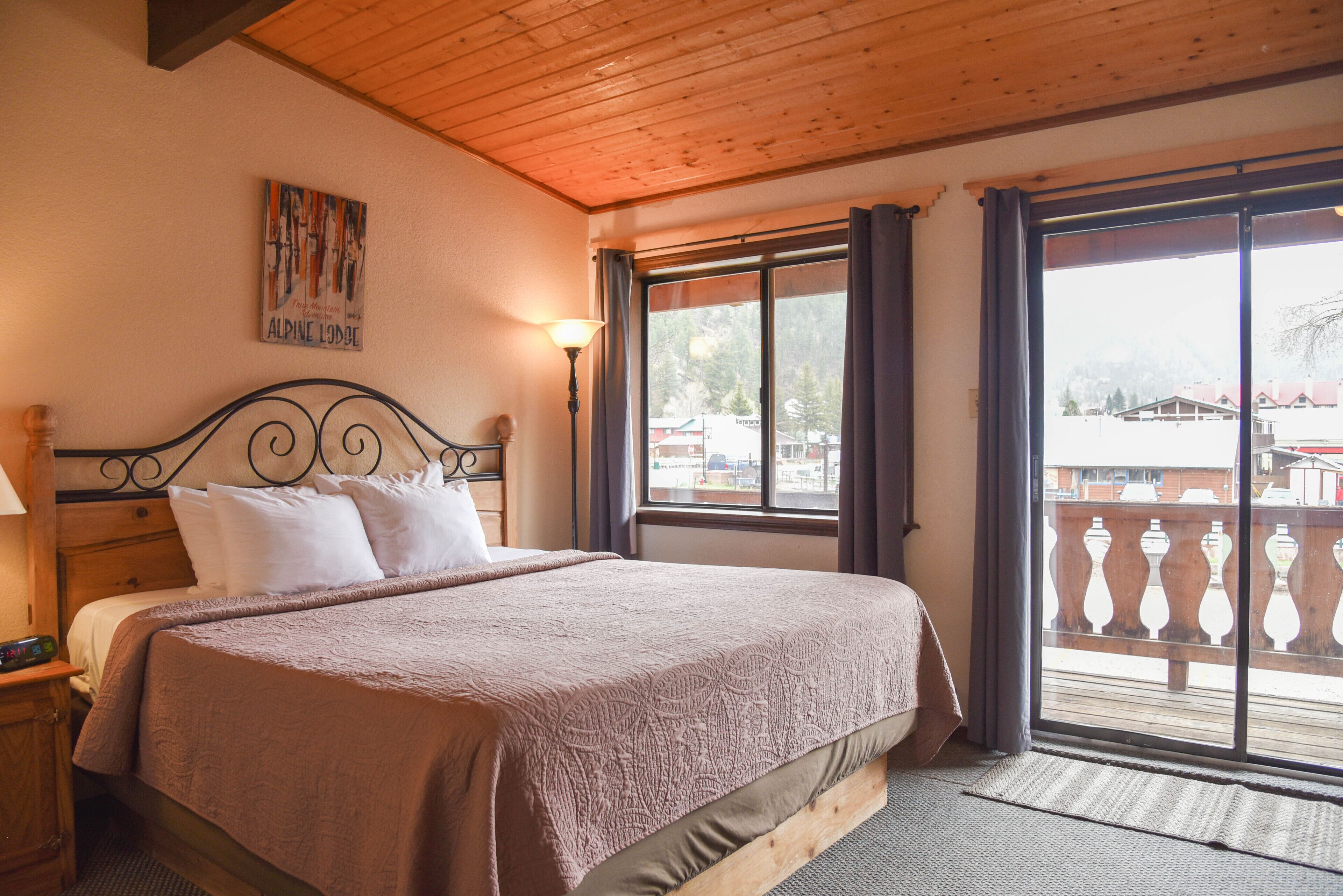 A bedroom with a wooden ceiling, a large bed with a headboard, a nightstand, a lamp, and a window with a view of buildings and trees.