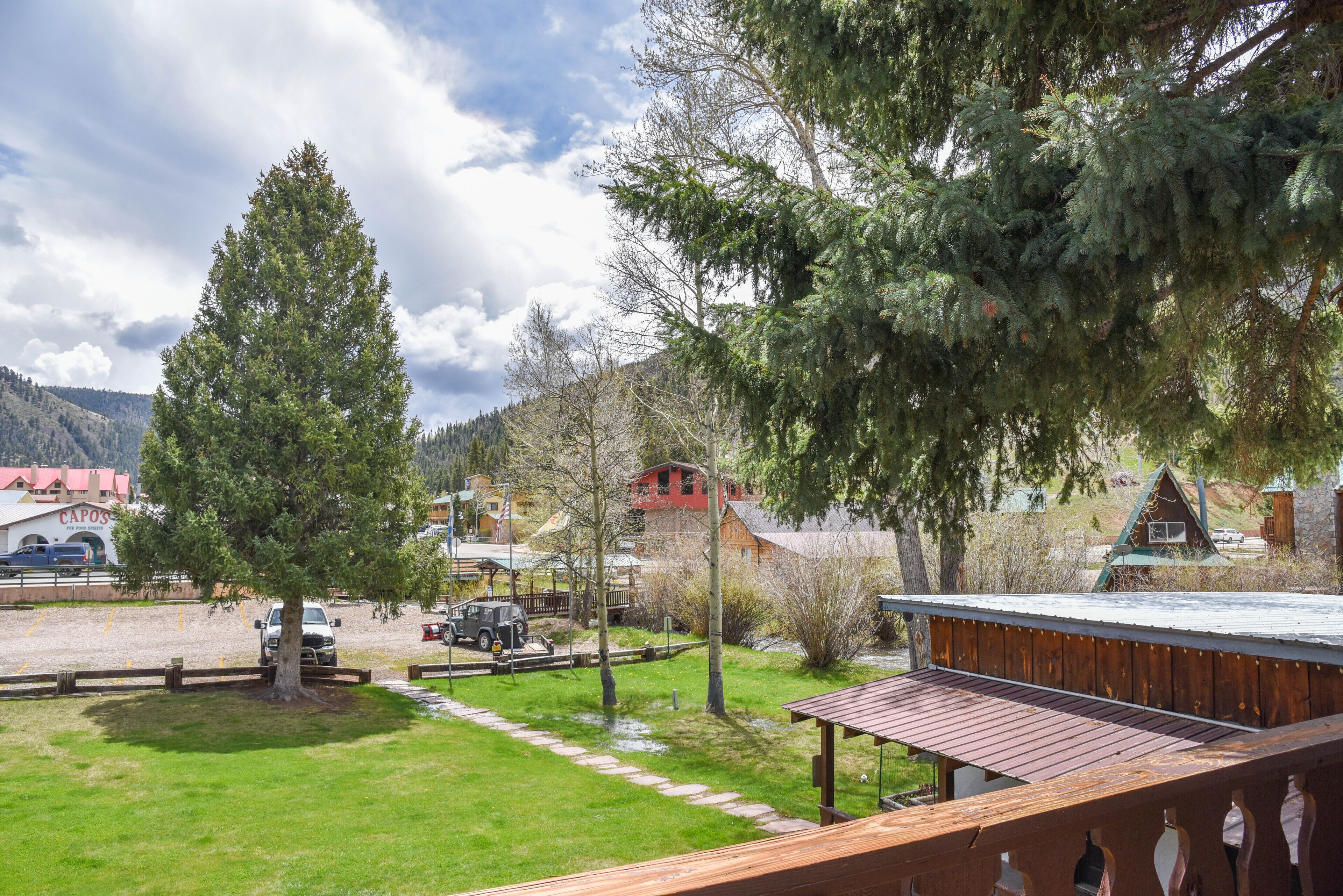 A wooden deck overlooking a grassy area with a large tree and a building with a red roof in the background.