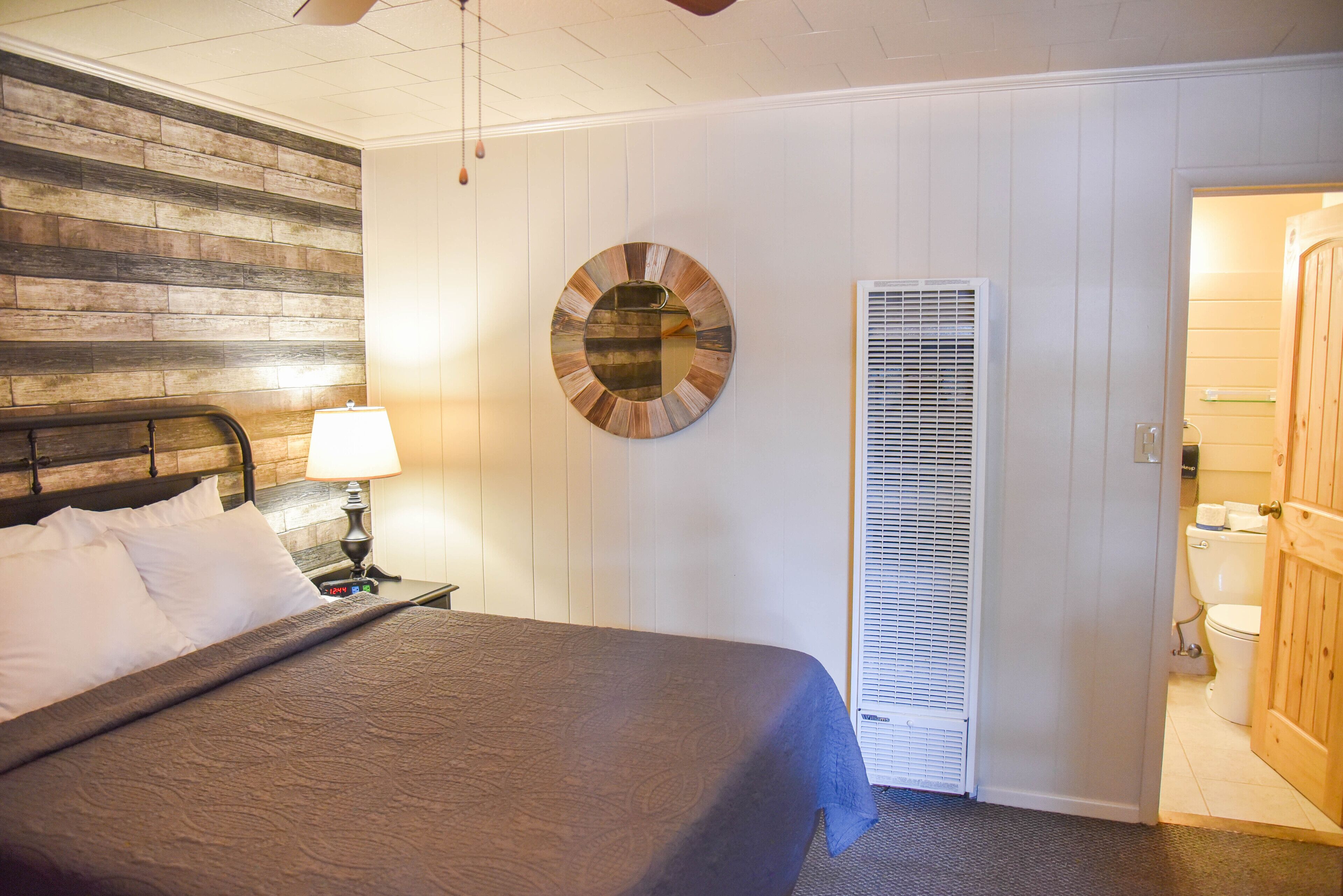 A bedroom with a bed, a nightstand with a lamp, a wooden headboard, a circular wall decoration, a window with blinds, and a wooden door leading to a bathroom.