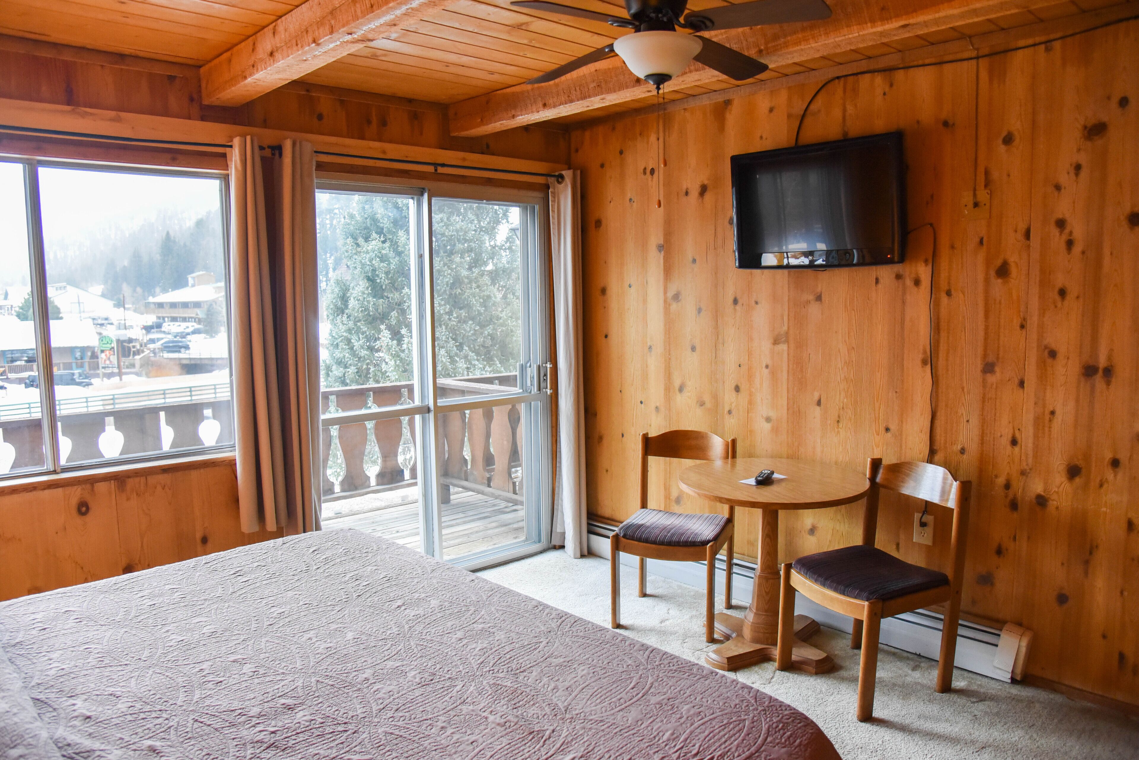 A wooden room with a bed, a table with two chairs, a television, and a large window with curtains.