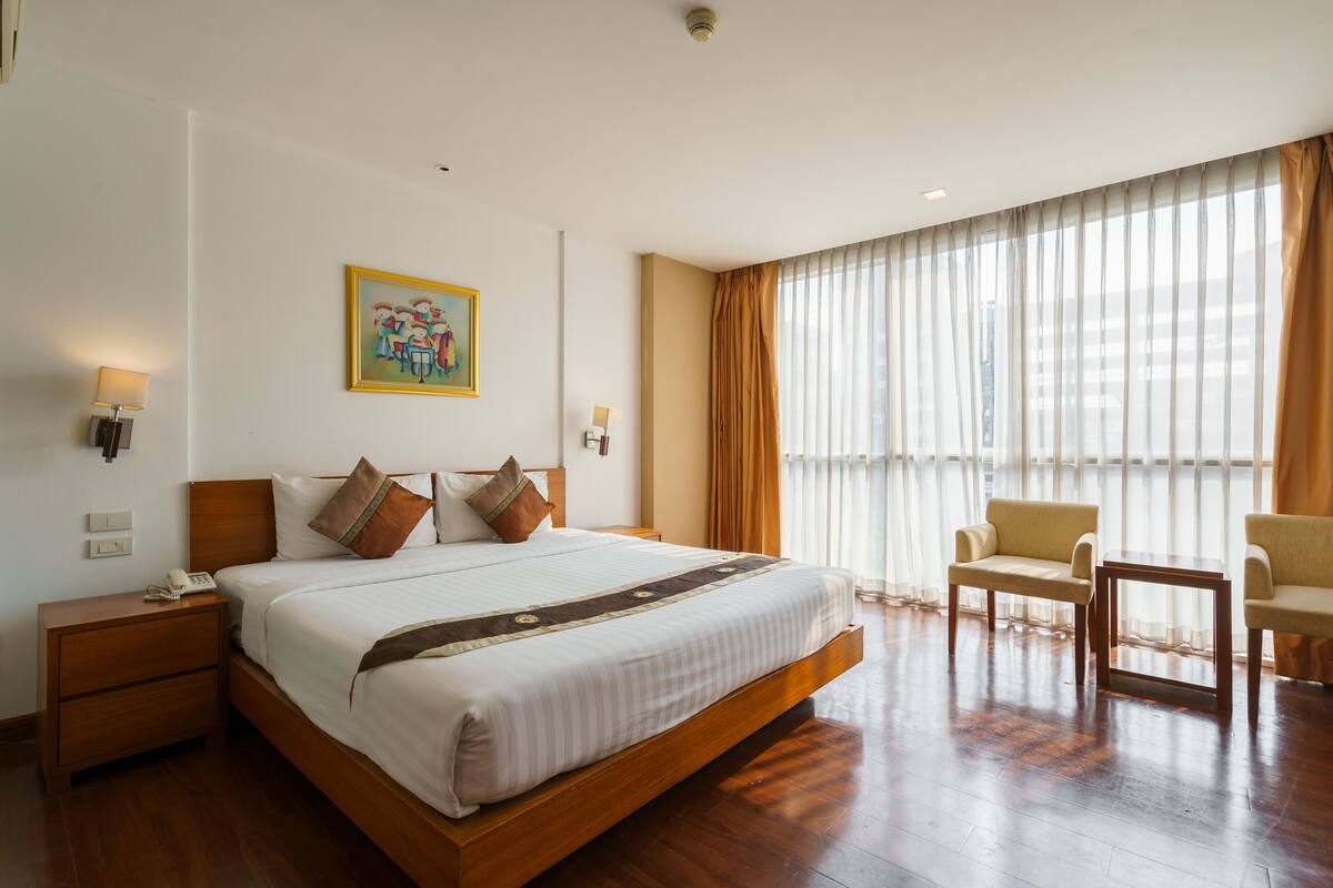 A hotel room with a large bed, bedside tables, a chair, and a view of the city through sheer curtains.