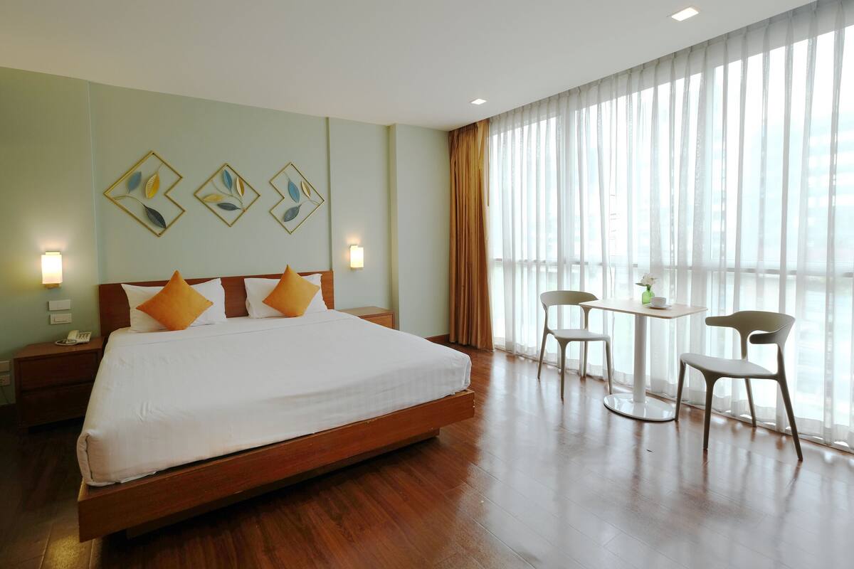 A hotel room with a large bed, two bedside tables with lamps, a small table with chairs, and a large window with curtains.