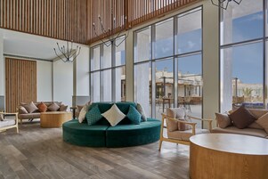 A spacious lobby with a green sectional sofa, wooden flooring, and large windows offering a view of the cityscape.