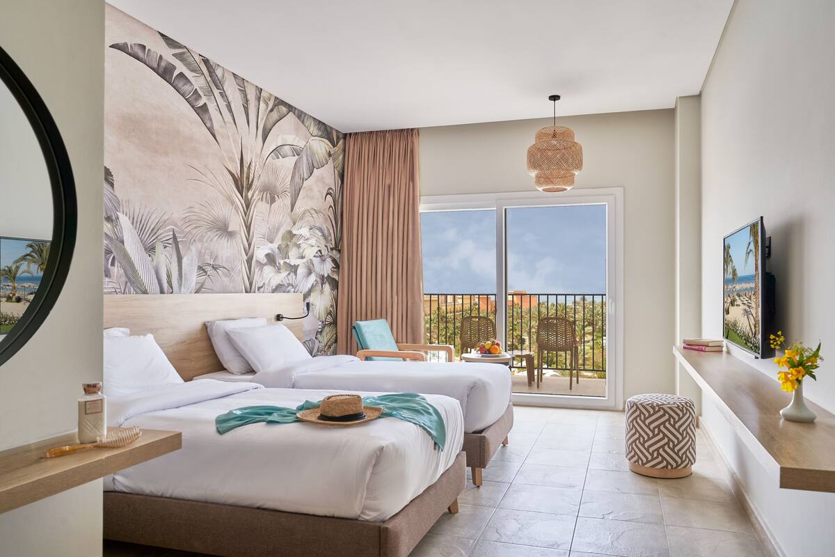 A hotel room with two beds, a large window, a balcony, and a painting on the wall.