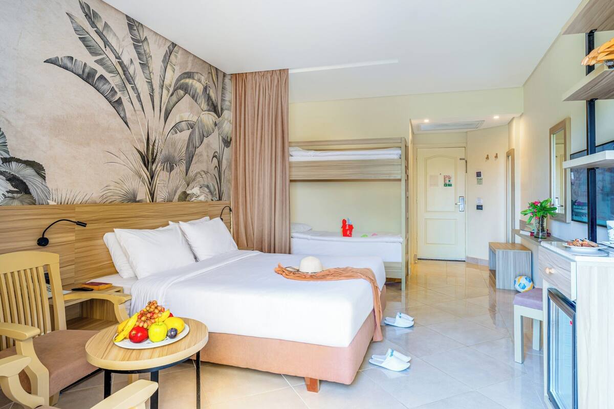 A hotel room with a bed, a desk, a chair, a small table with fruit, and a wall with a plant mural.