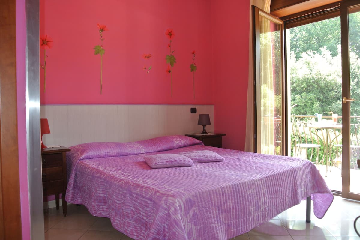 Un dormitorio con cama, mesita de noche, lámpara y ventana con vista a follaje.