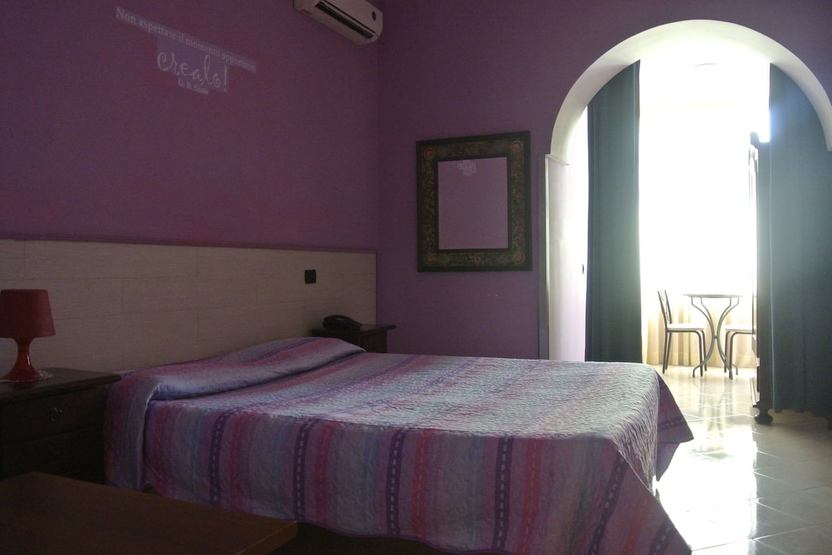 Un dormitorio con cama, mesita de noche, lámpara, espejo y vistas al exterior desde una puerta abierta.