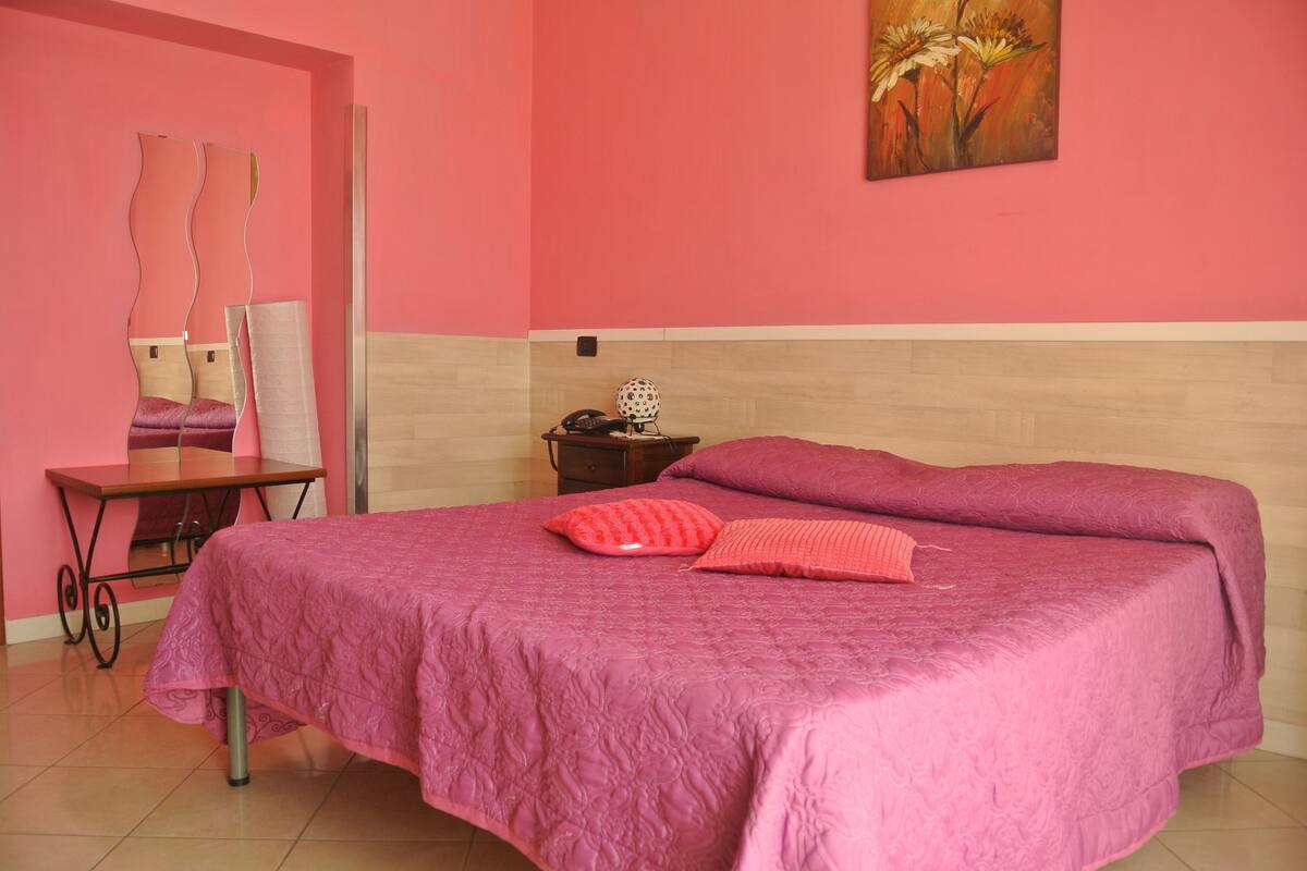Un dormitorio con una colcha rosa, una mesita de noche con lámpara, un espejo y un cuadro en la pared.