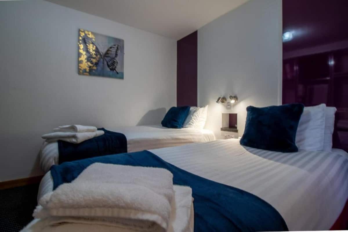 A hotel room with two beds, each with white linens and blue pillows. There is a painting on the wall and a nightstand with a lamp.
