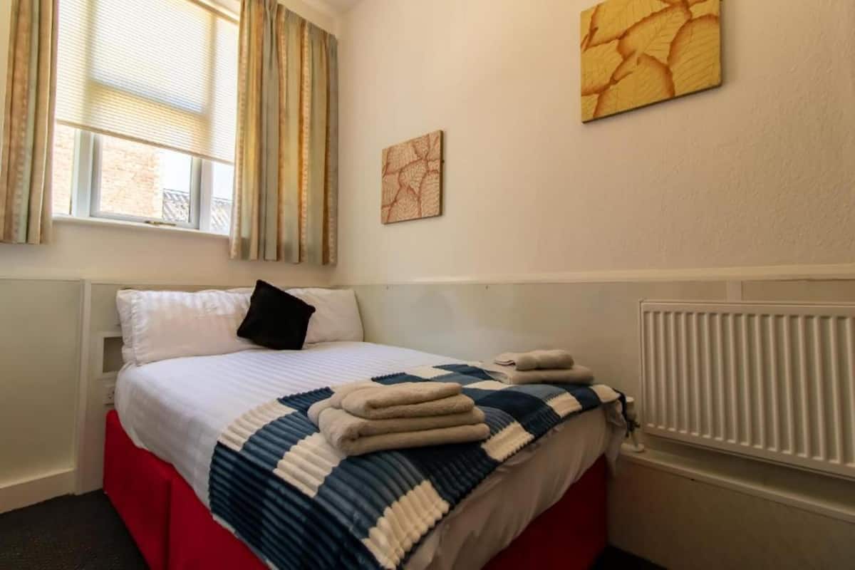 A hotel room with a bed, a window with curtains, a radiator, and two wall art pieces.