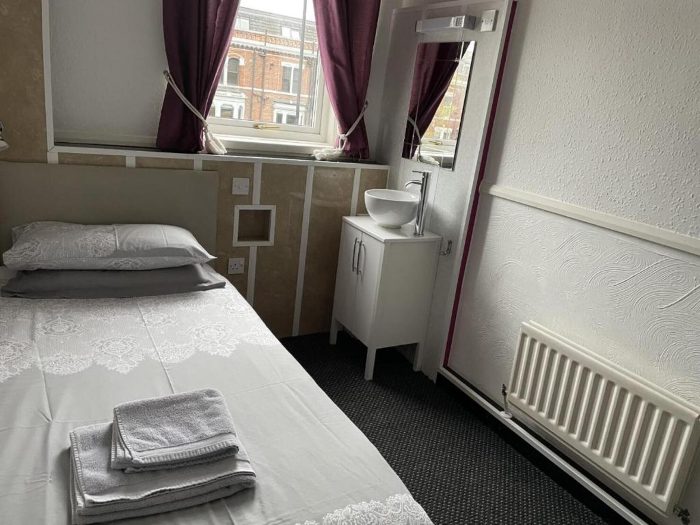 A hotel room with a bed, a small white cabinet with a sink, and a window with curtains.