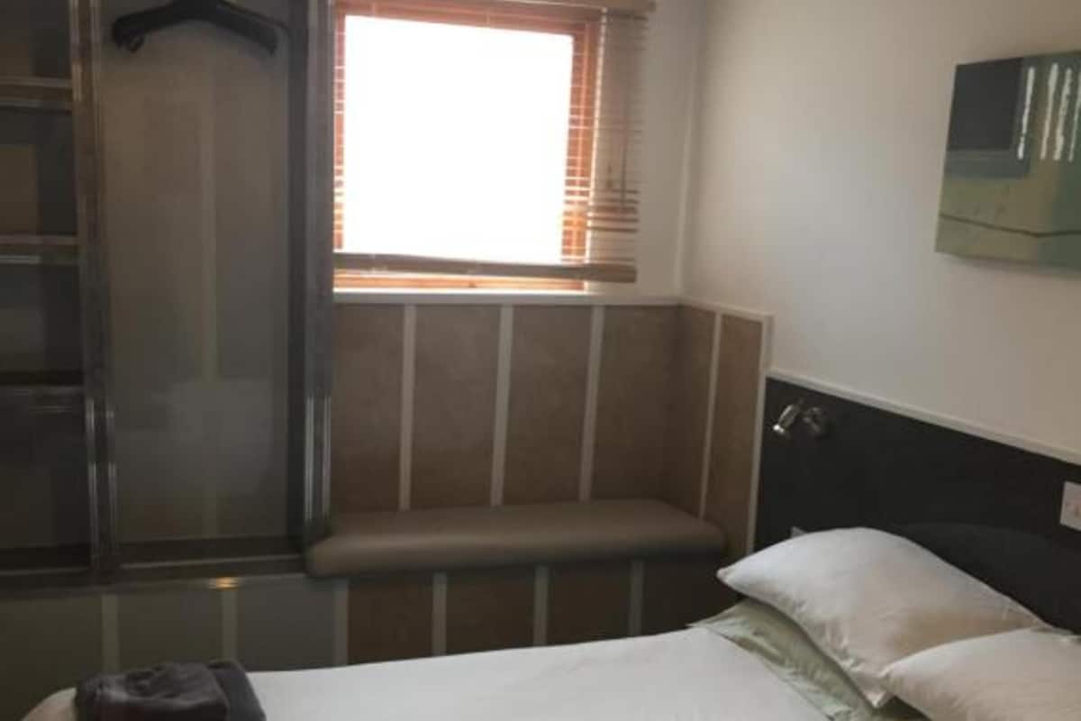 A hotel room with a bed, a bench, a hanging coat rack, and a window with blinds.