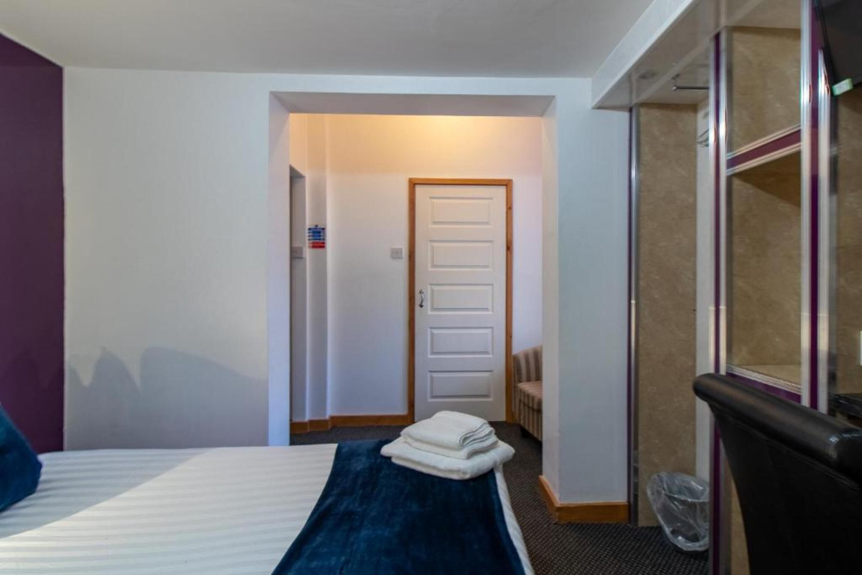 A hotel room with a bed, a blue blanket, a white door, and a trash bin.