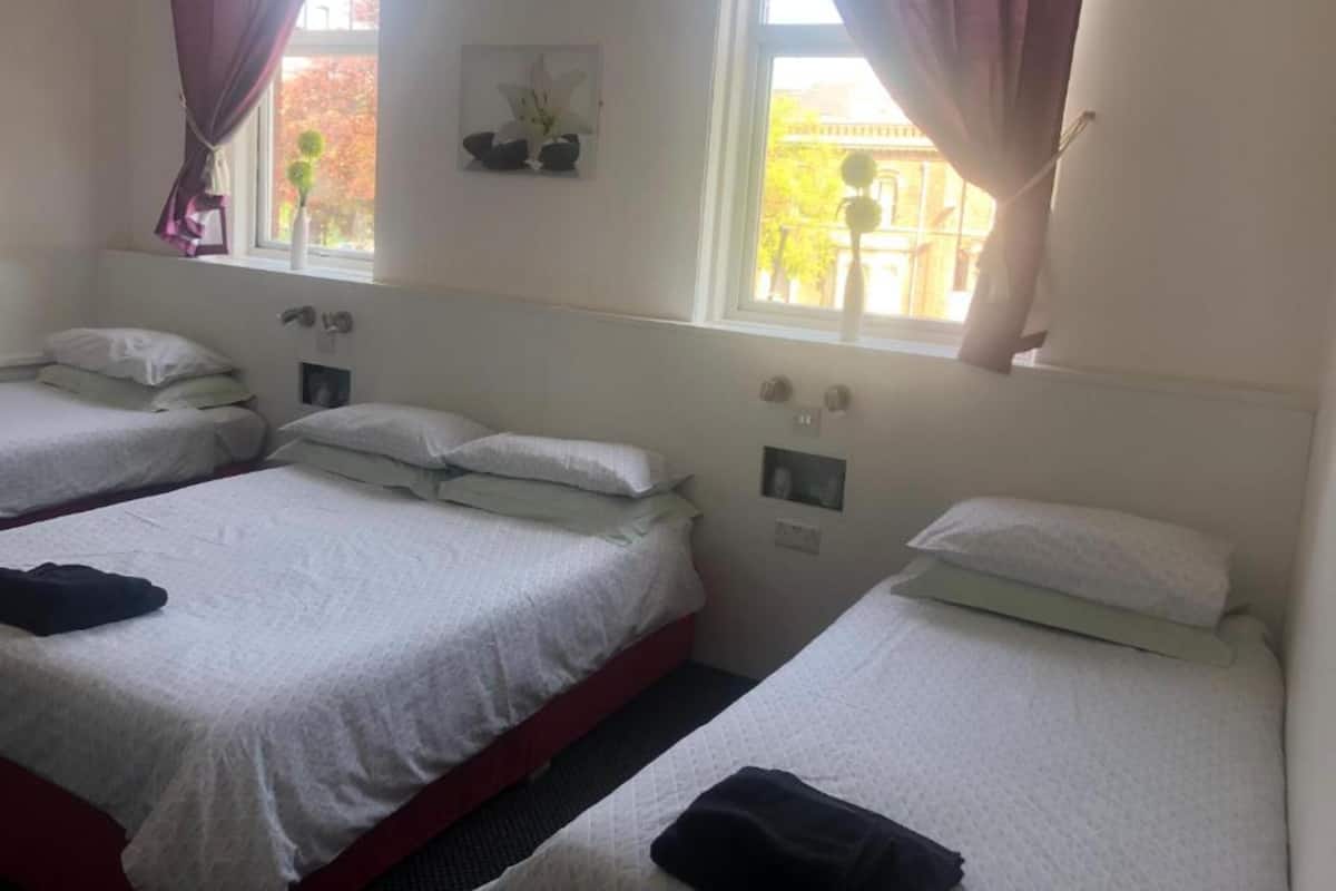 A hotel room with three single beds, each with white bedding and pillows. There are two windows with curtains, and a small picture frame on the wall.