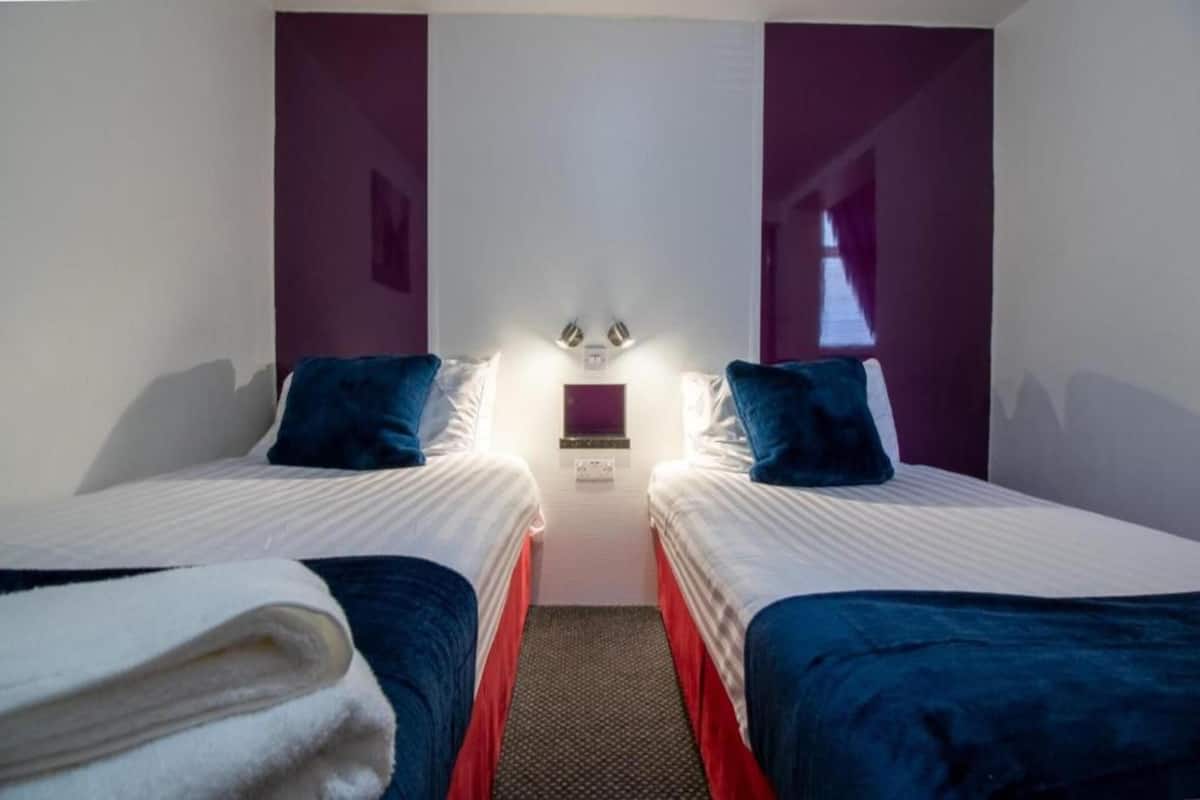 A hotel room with two beds, each with a blue headboard, white bedding, and a red accent. There is a small table between the beds, and a wall-mounted light fixture above it.
