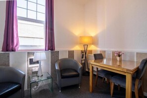 Habitación de hotel con una mesa de vidrio, dos sillones, una lámpara y vista al mar a través de una ventana con cortinas moradas.