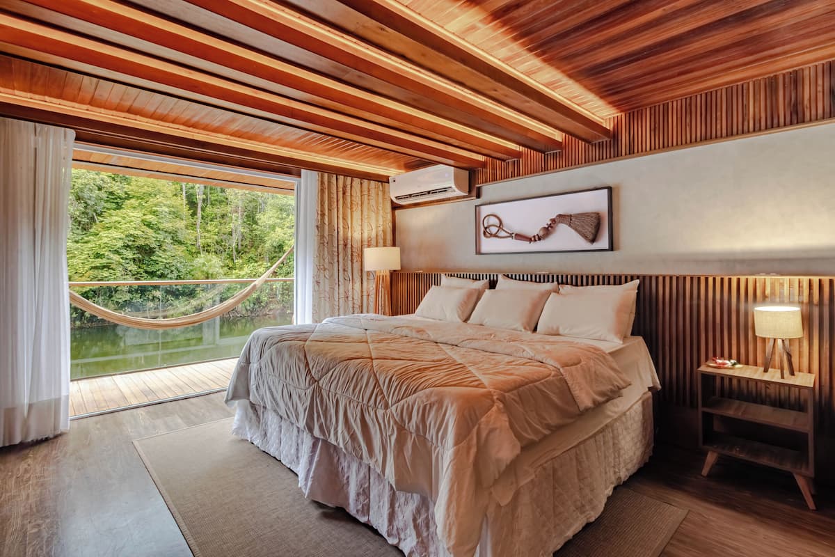 A bedroom with a large bed, wooden ceiling, a hammock, a nightstand with a lamp, and a view of greenery outside.