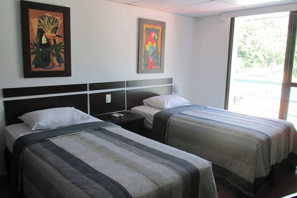 A hotel room with two single beds, a nightstand, a wall-mounted TV, and two framed artworks.