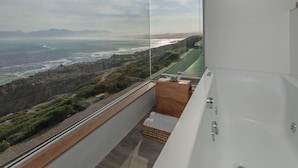 Une salle de bain moderne avec une grande baignoire, un balcon vitré offrant une vue sur l’océan et un banc en bois.