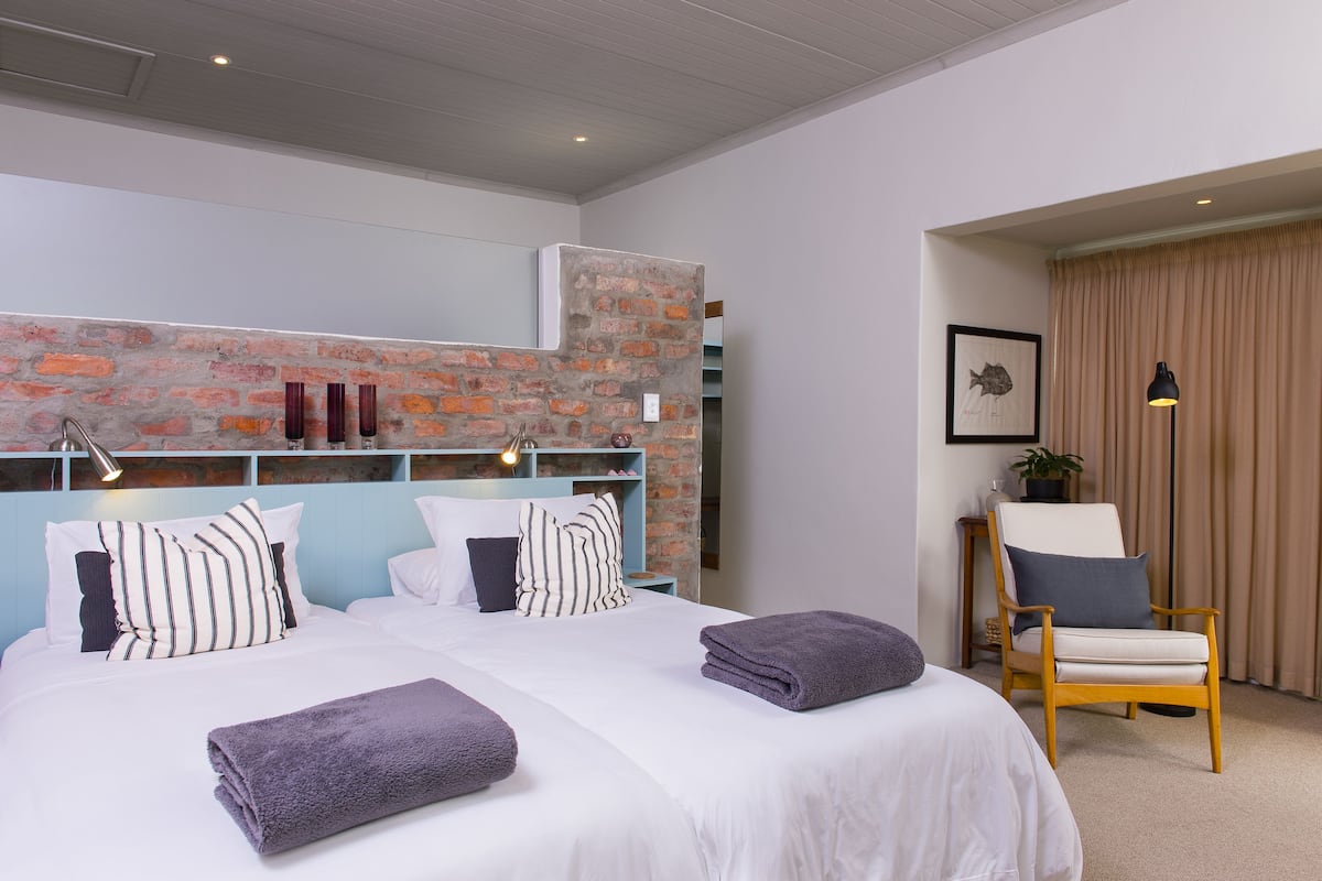 Habitación de hotel con pared de ladrillo decorativa, dos camas con ropa blanca, una silla de madera y un cuadro en la pared.