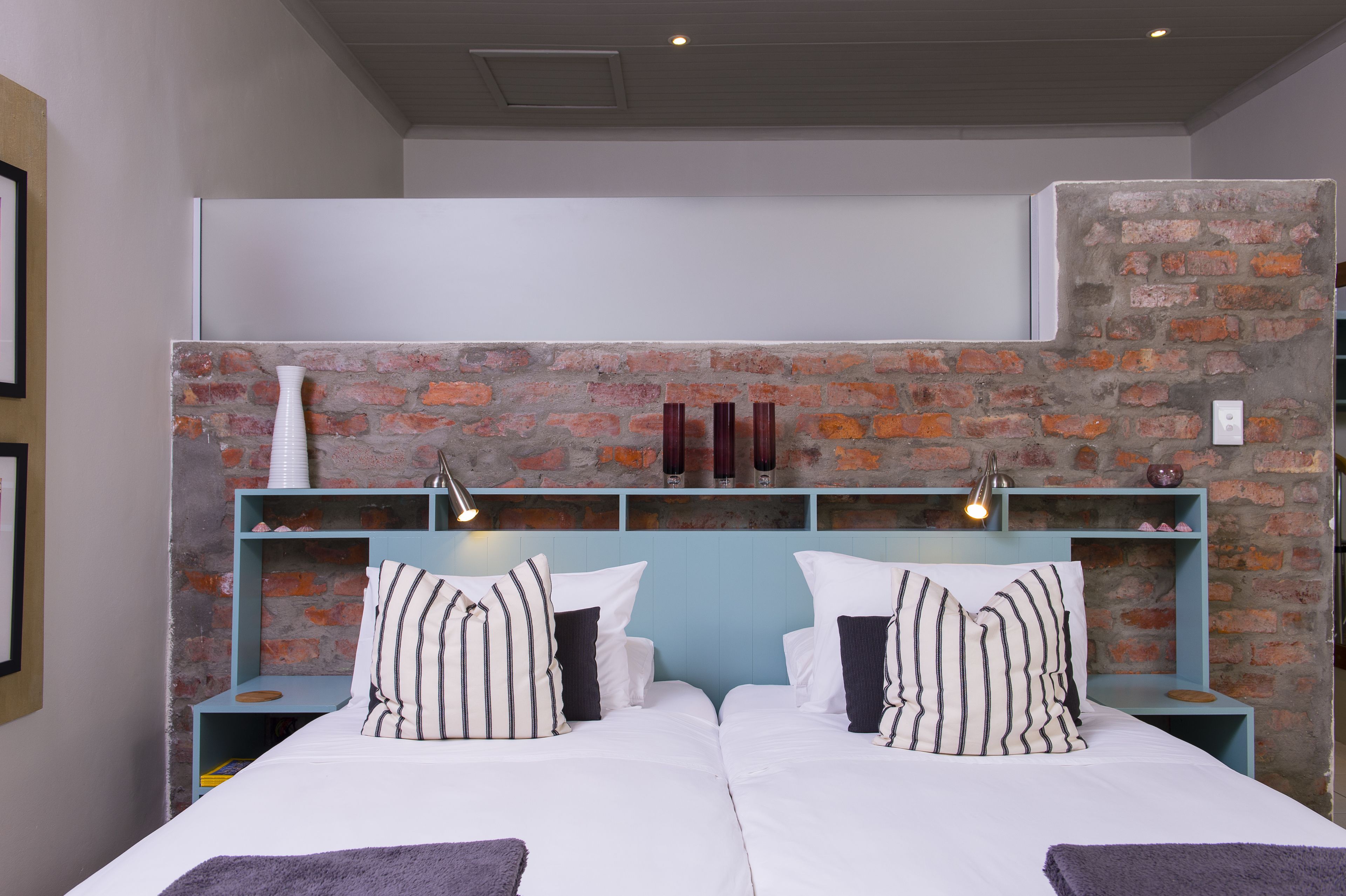 A double bed with white bedding and striped pillows, a brick accent wall, and a blue nightstand with a lamp.