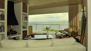 Un salon moderne avec un canapé blanc, une table basse et une bibliothèque. Une grande fenêtre offre une vue sur la mer.