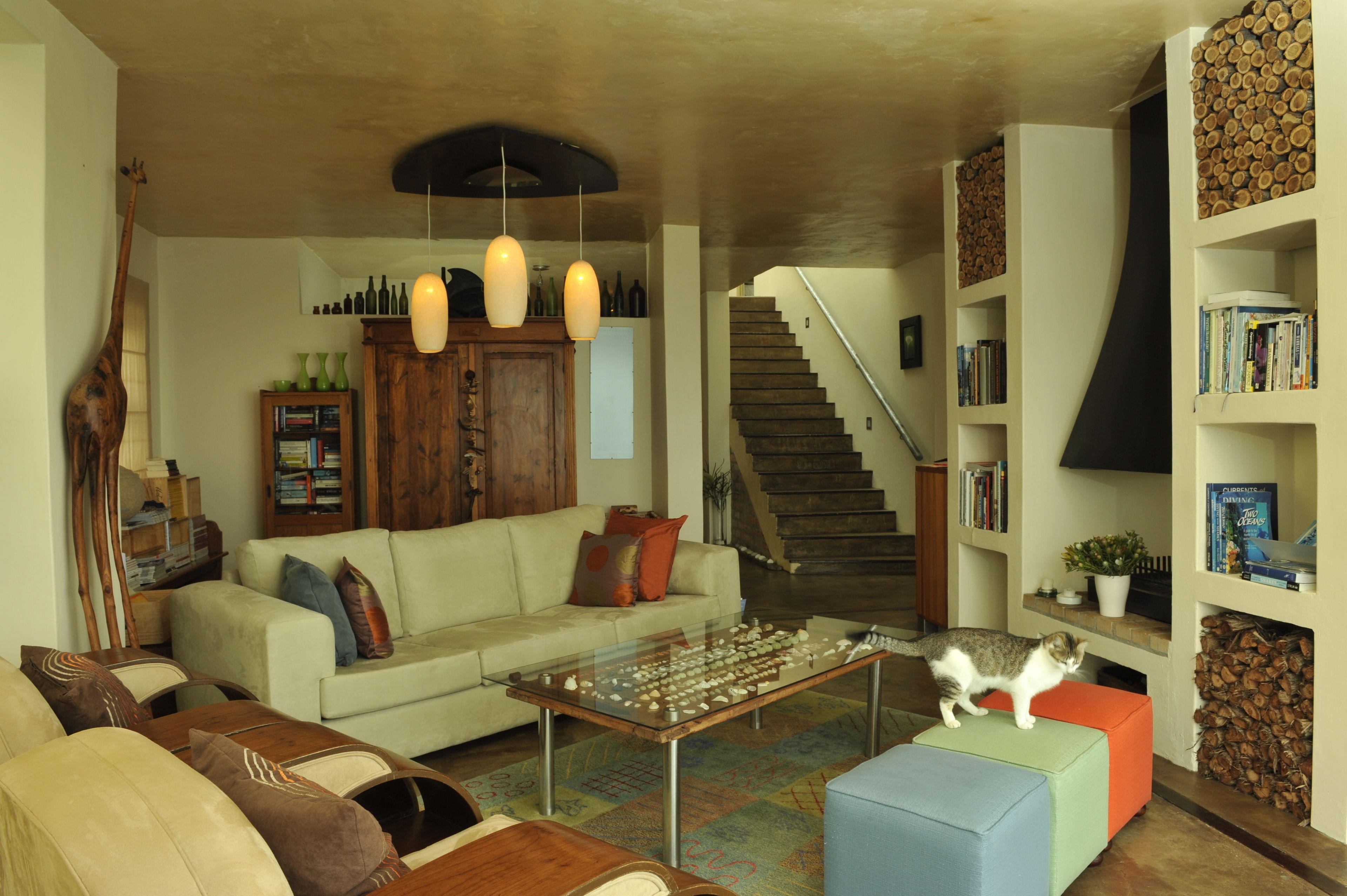 A living room with a sofa, coffee table, and a cat on an ottoman. There is a staircase and bookshelves with books and decorative items.