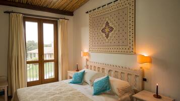 A bedroom with a large bed, two bedside lamps, a wall tapestry, and a view through a glass door.