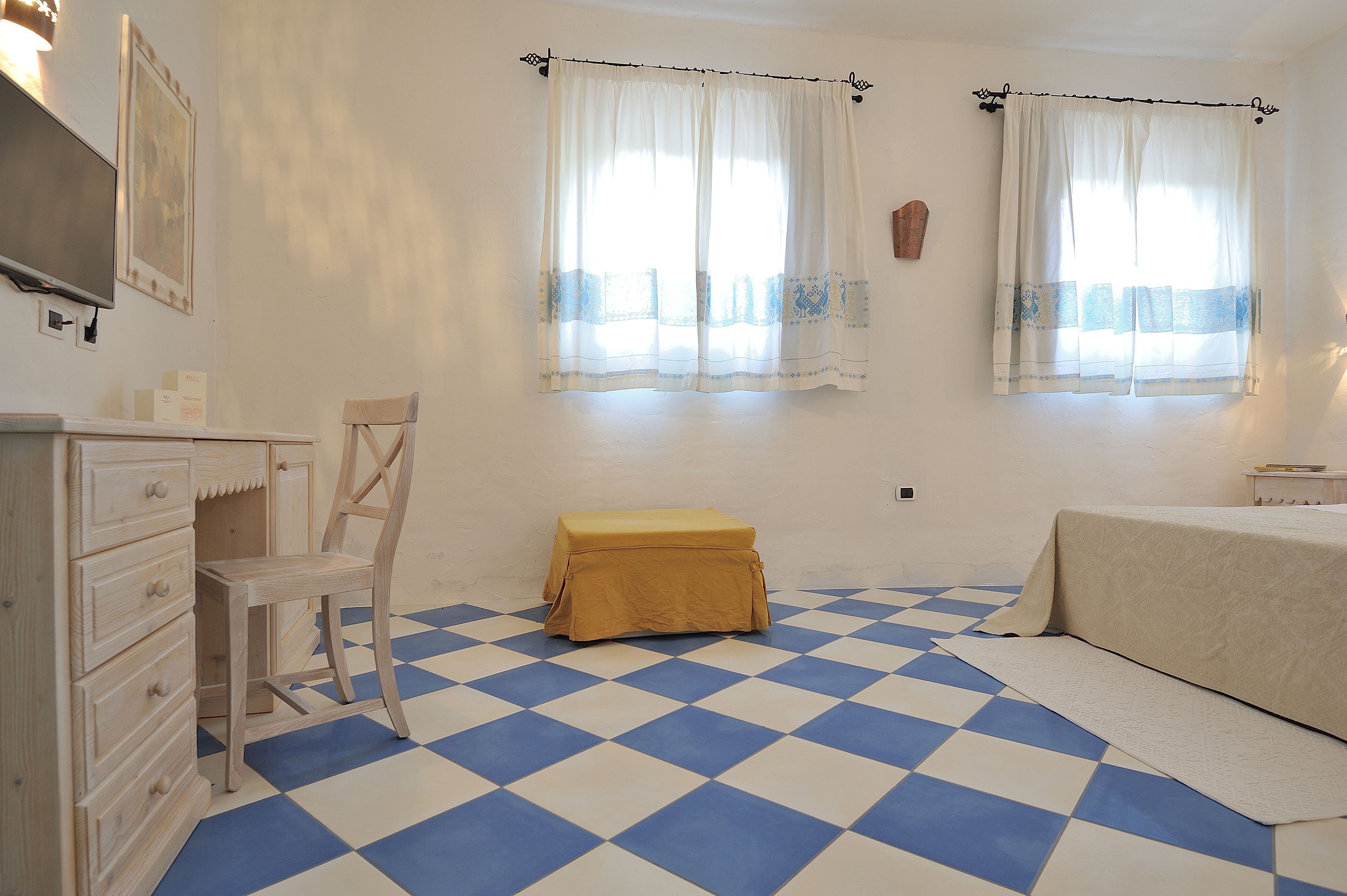 A room with a checkered floor, a wooden chair, a bed with a beige cover, and two windows with sheer curtains.