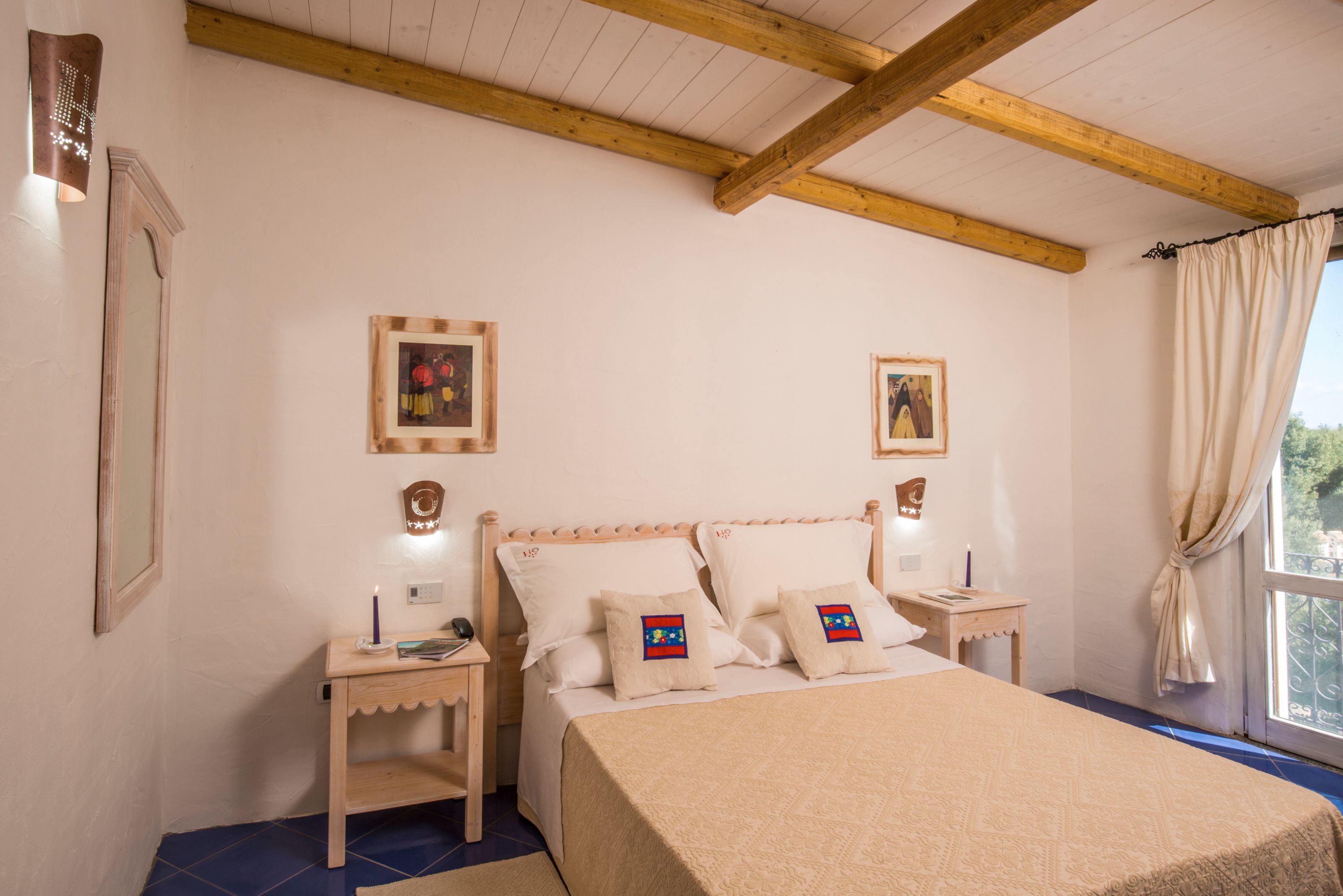 A bedroom with a wooden ceiling, two bedside tables, a bed with a beige quilt, and two framed pictures on the wall.