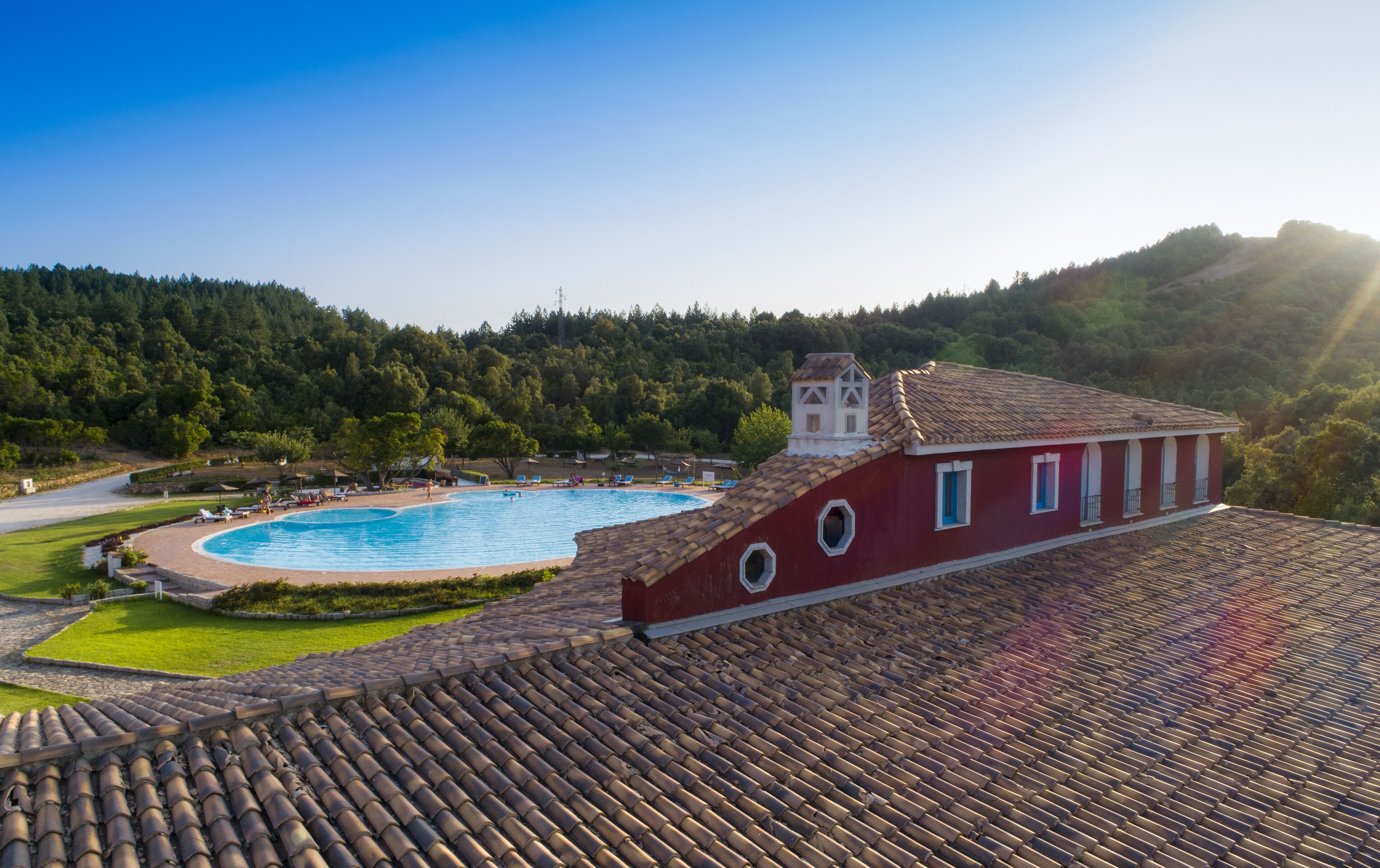 Un edificio rojo con techo de tejas, piscina y una ladera boscosa al fondo.