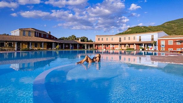 A swimming pool with a person relaxing in the water, surrounded by buildings with a Mediterranean architectural style.