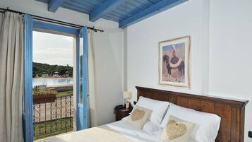 Ein Schlafzimmer mit hölzernem Kopfteil, einem Bett mit weißen Laken und herzförmigen Kissen, einem Nachttisch mit Lampe sowie Blick auf einen Pool und Grünanlagen durch eine offene Tür.