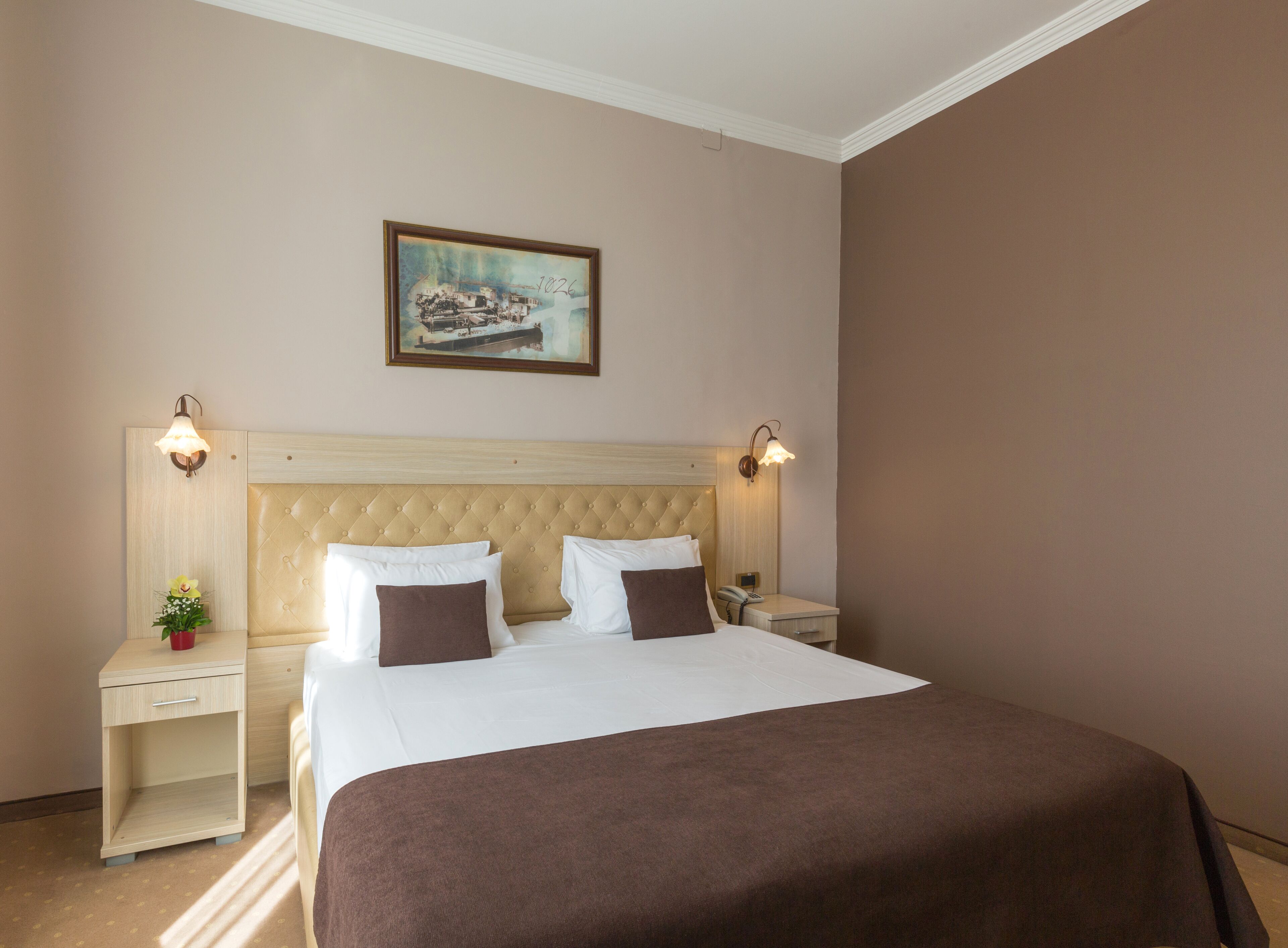 Habitación de hotel con una cama grande, dos mesitas de noche, una lámpara auxiliar, un cuadro colgado en la pared y un jarrón decorativo.