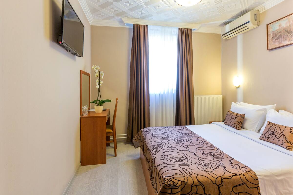 Habitación de hotel con cama, escritorio, silla, televisor y ventana con cortinas.