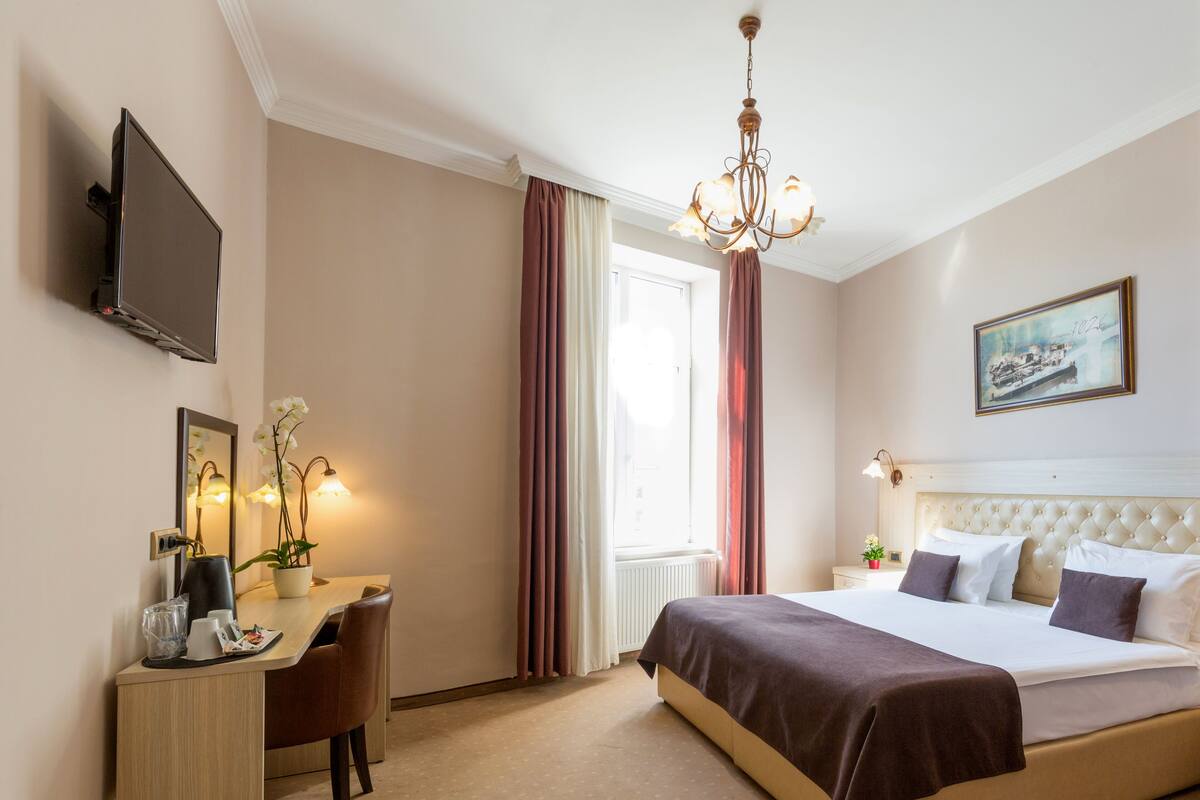 Habitación de hotel con cama, escritorio, silla, televisor, candelabro y ventana con cortinas.