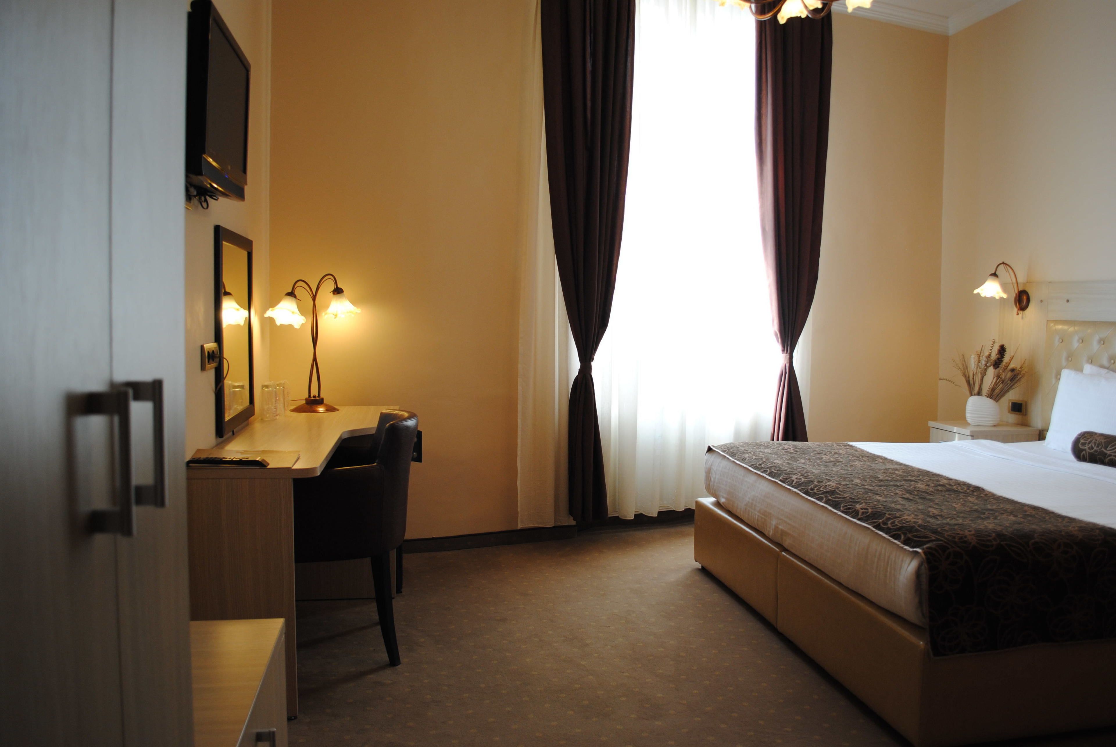 A hotel room with a bed, a desk, and a chair. There is a window with curtains, a lamp, and a mirror on the wall.