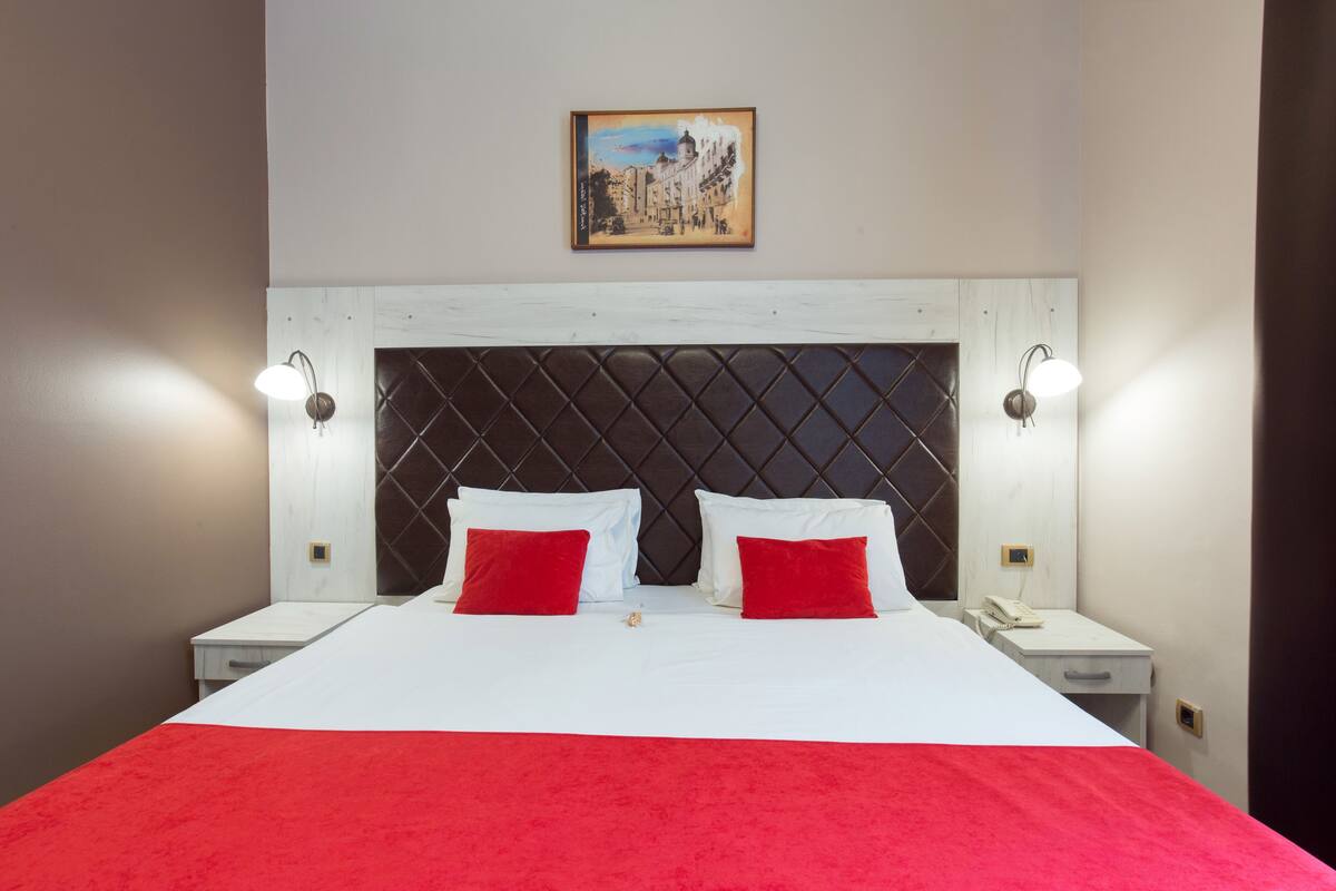 Una habitación de hotel con una cama grande, dos almohadas rojas, una colcha blanca, un cabecero acolchado, dos mesitas de noche con lámparas y un cuadro en la pared.