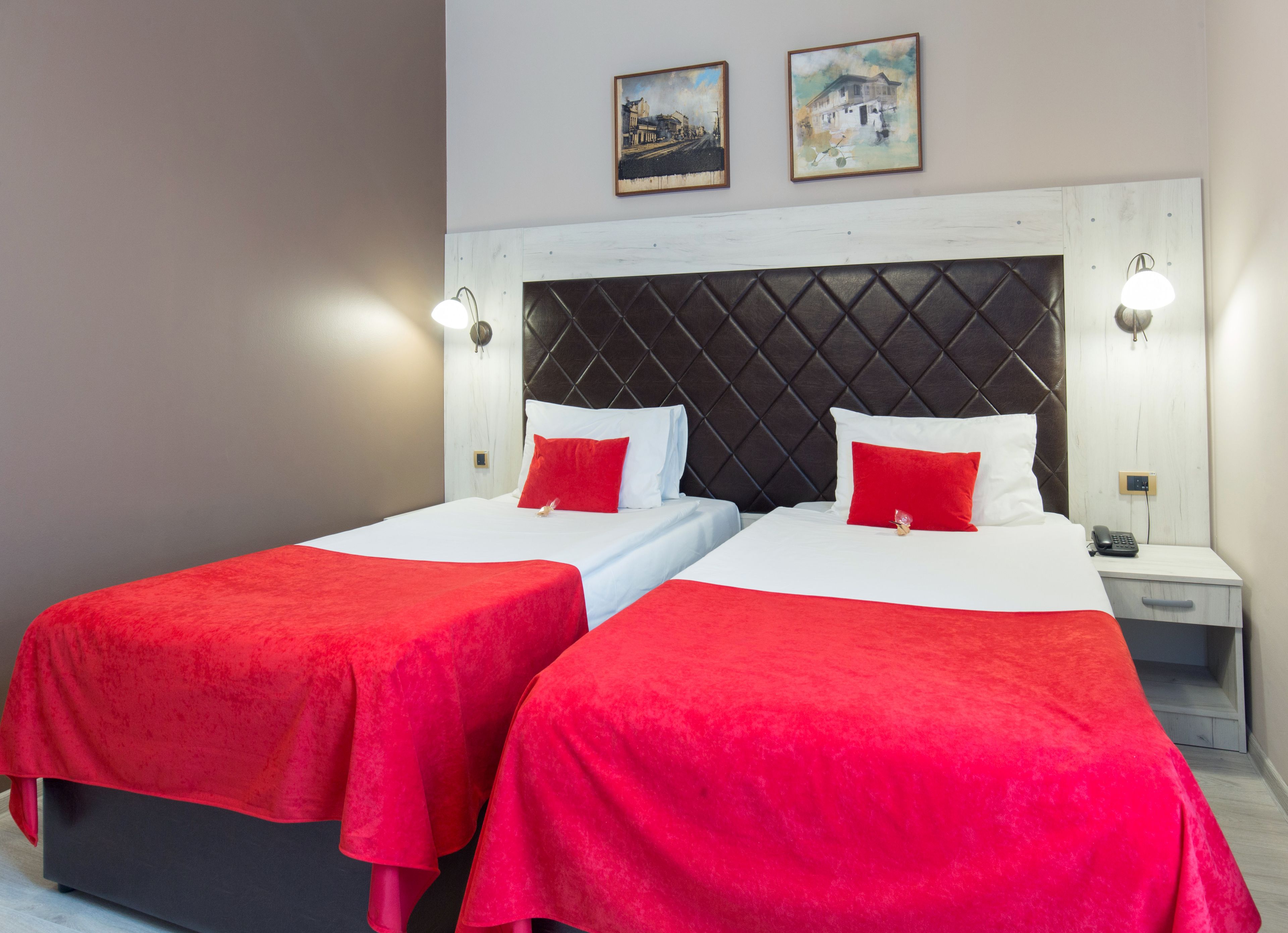 Een hotelkamer met twee bedden, elk voorzien van een rode sprei en witte lakens. Twee wandlampen staan op nachtkastjes, en de kamer heeft een wit hoofdeinde met een ruitpatroon.