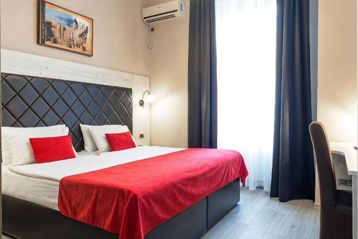 Una habitación de hotel con cama, escritorio, silla, un cuadro en la pared y ventana con cortinas.