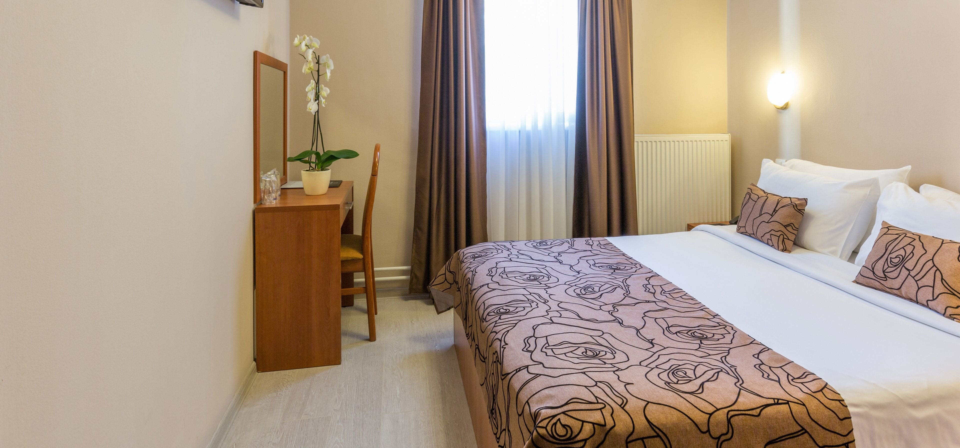 Hotellihuone, jossa on sänky, työpöytä, tuoli, ruukkukasvi ja ikkuna, jossa on verhot.