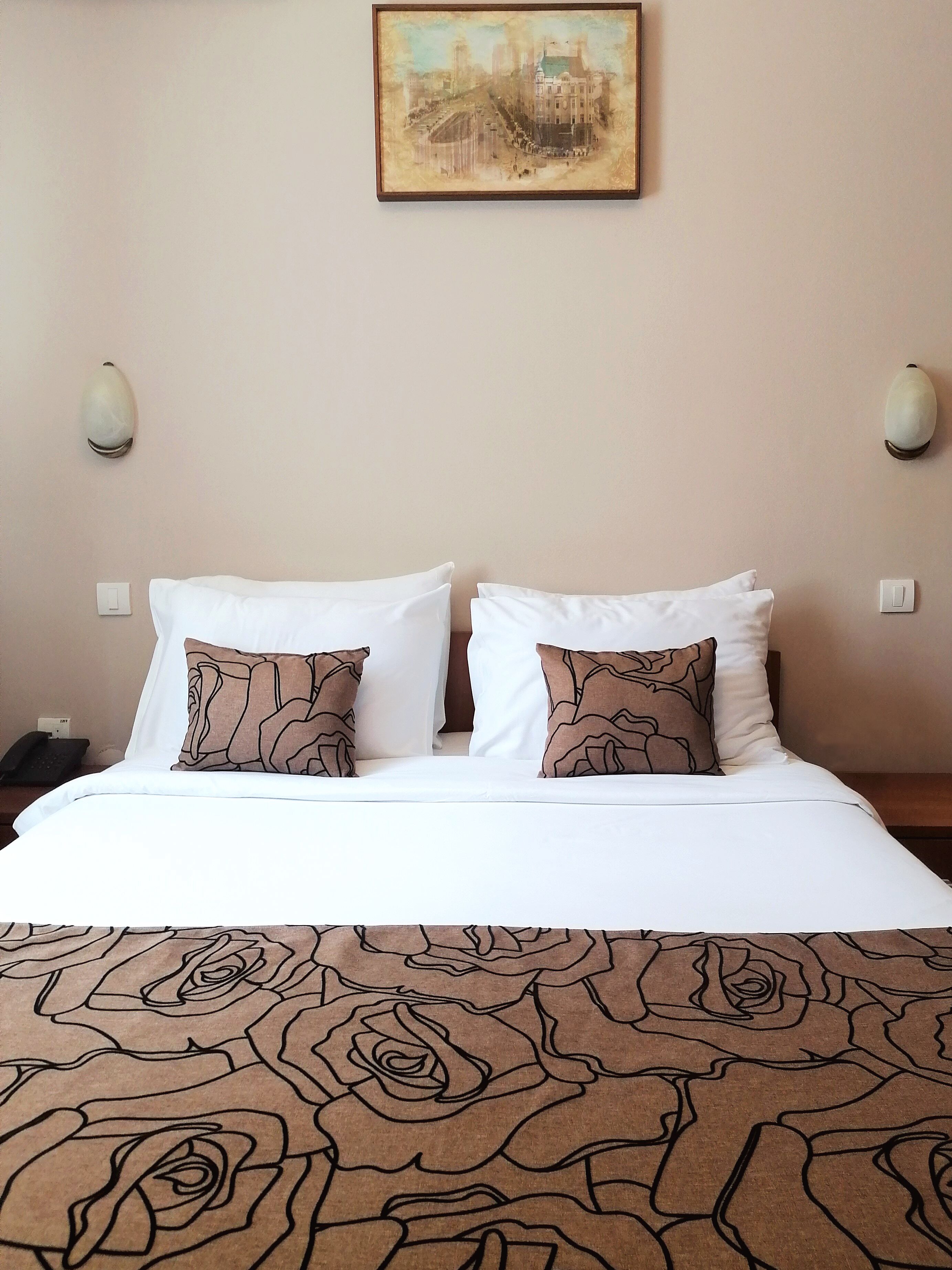 A hotel room with a bed, two wall sconces, a painting above the bed, and a telephone on the nightstand.