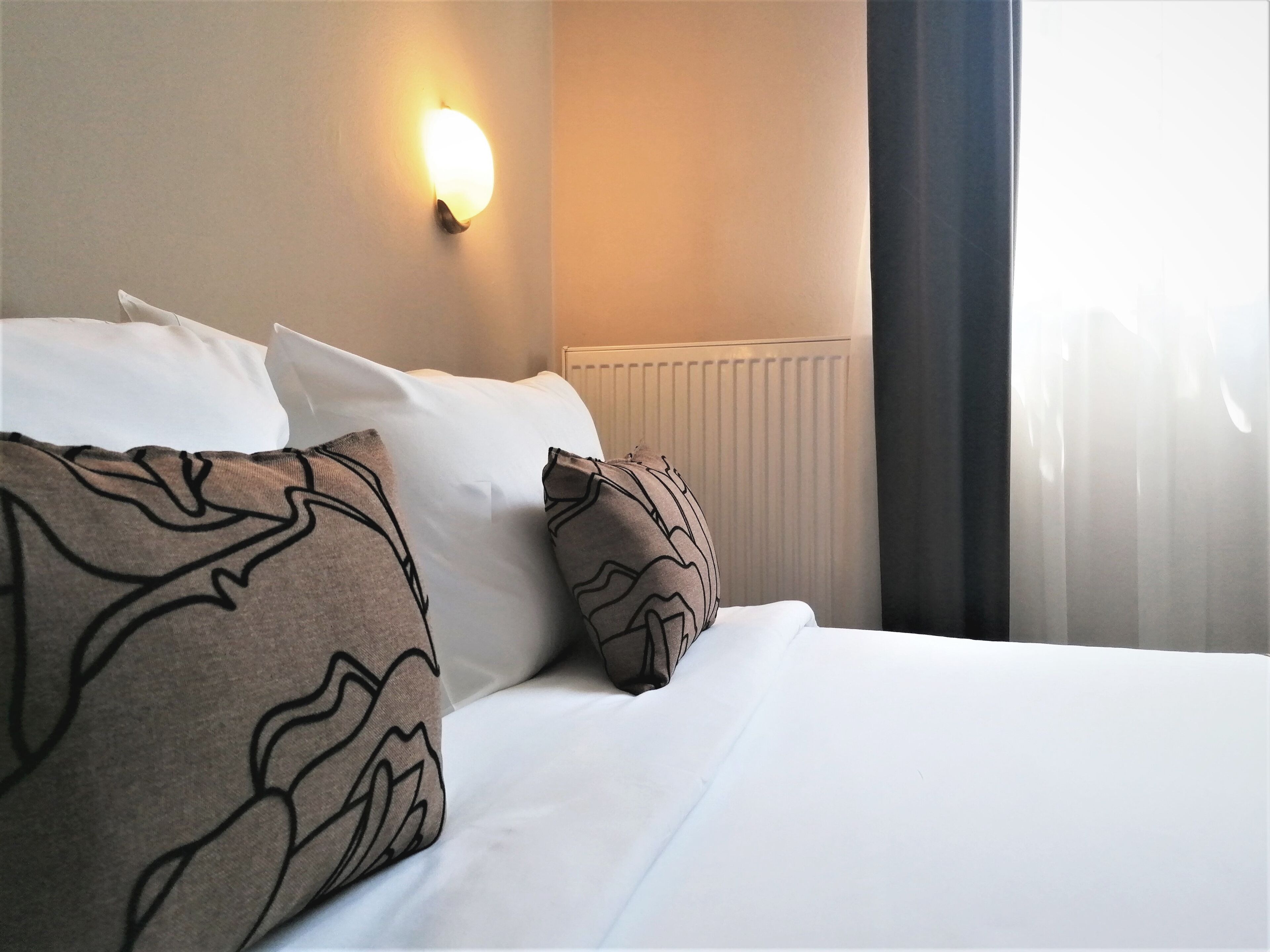Ein Hotelzimmer mit Bett, Kissen, einer Wandlampe und einem Fenster mit Vorhängen.
