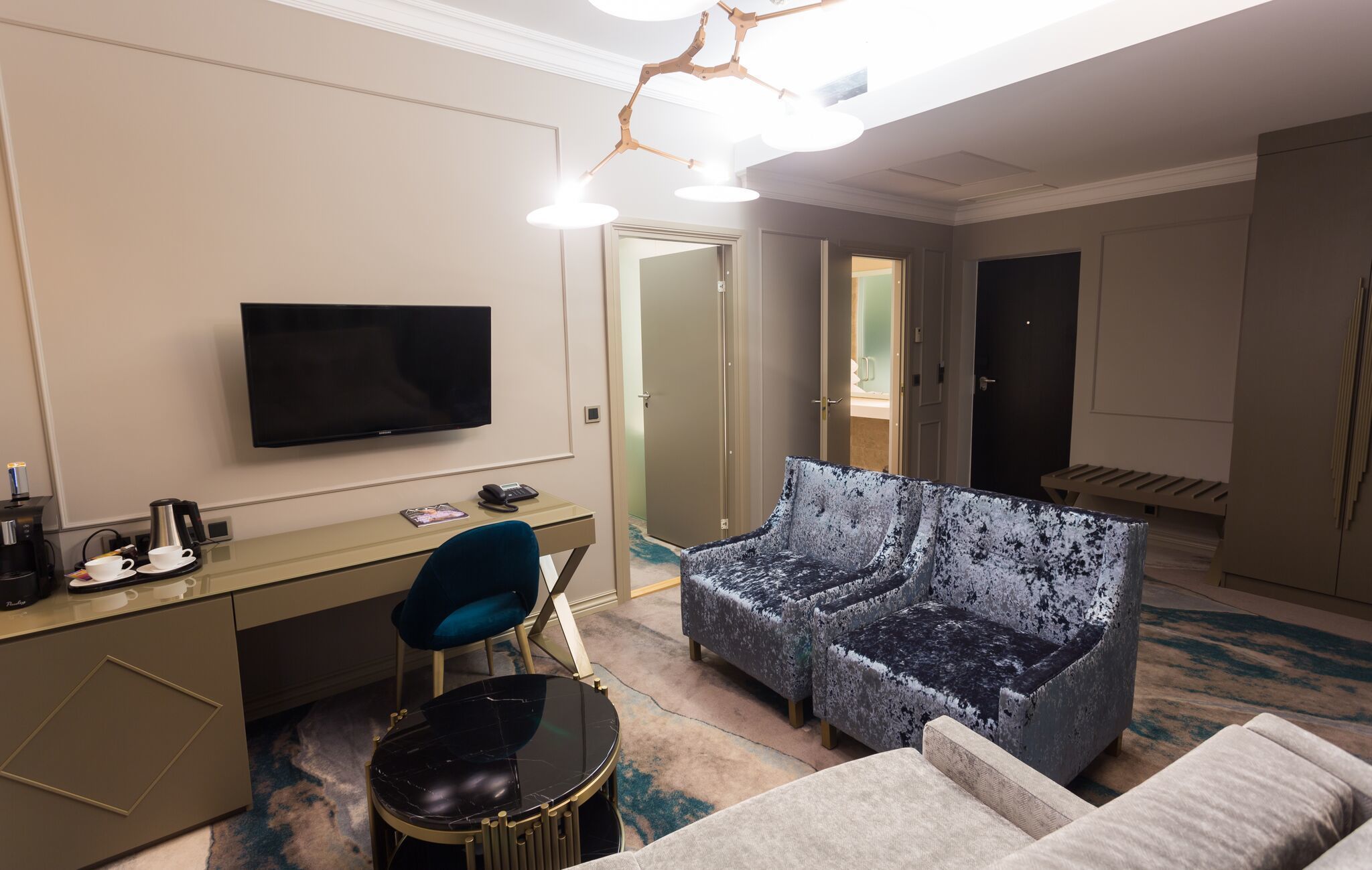 A hotel room with a flat-screen TV, a desk with a coffee maker, a blue chair, a patterned sofa, and a small table.